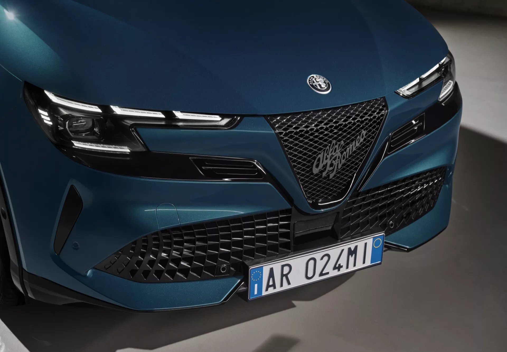 Zbliżony widok na przednią część Alfa-Romeo Junior 2024 skupiający się na grillu, emblemacie i lampach LED z eleganckim i agresywnym designem, wykonany z niskiego kąta po lewej stronie