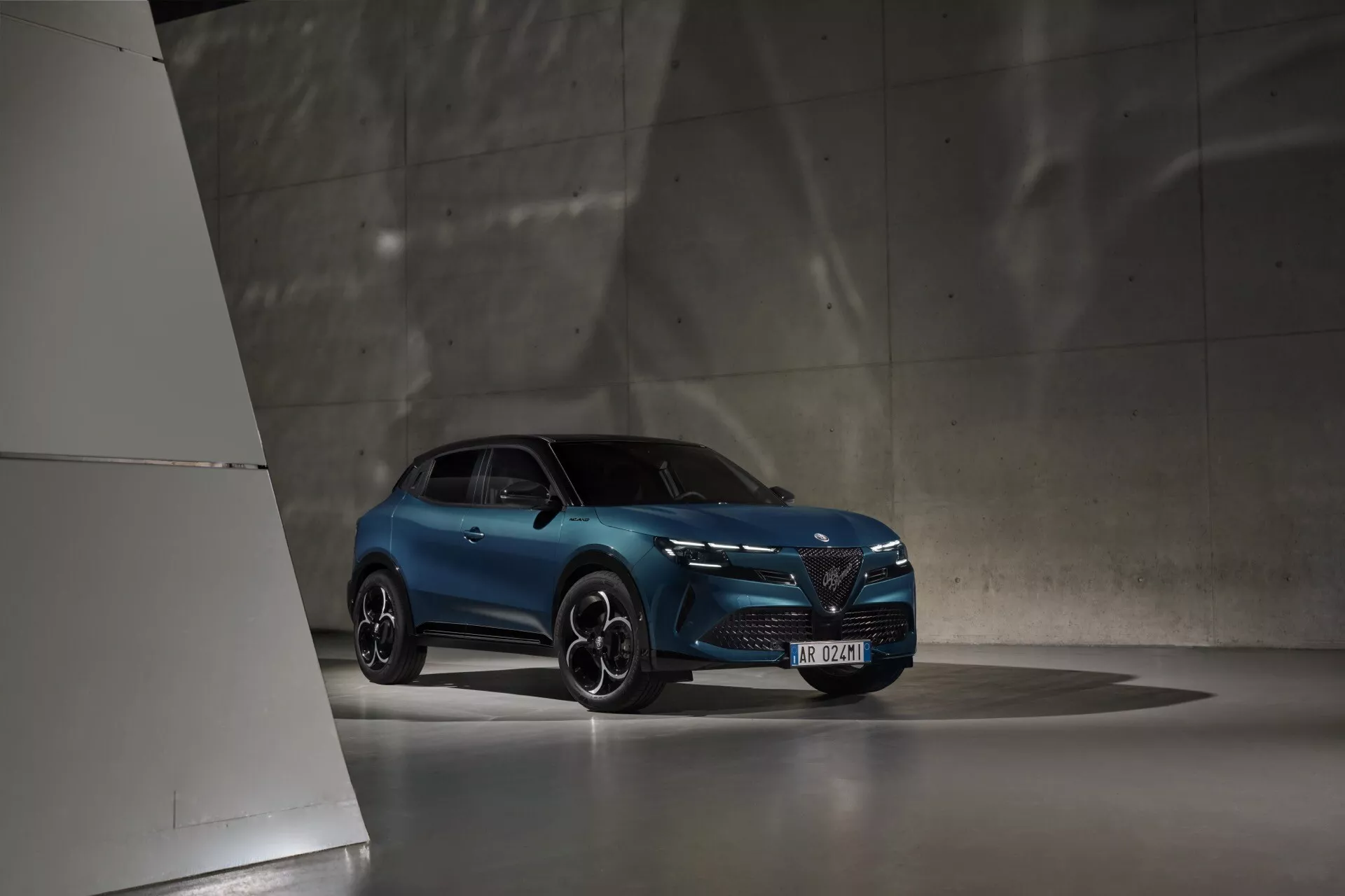 Niebieski Alfa Romeo Junior 2024 w słabo oświetlonym wnętrzu, zdjęcie z niskiego kąta podkreślające nowoczesny design i sportową postawę, z reflektorami LED, charakterystyczną przednią kratą i sportowymi felgami.