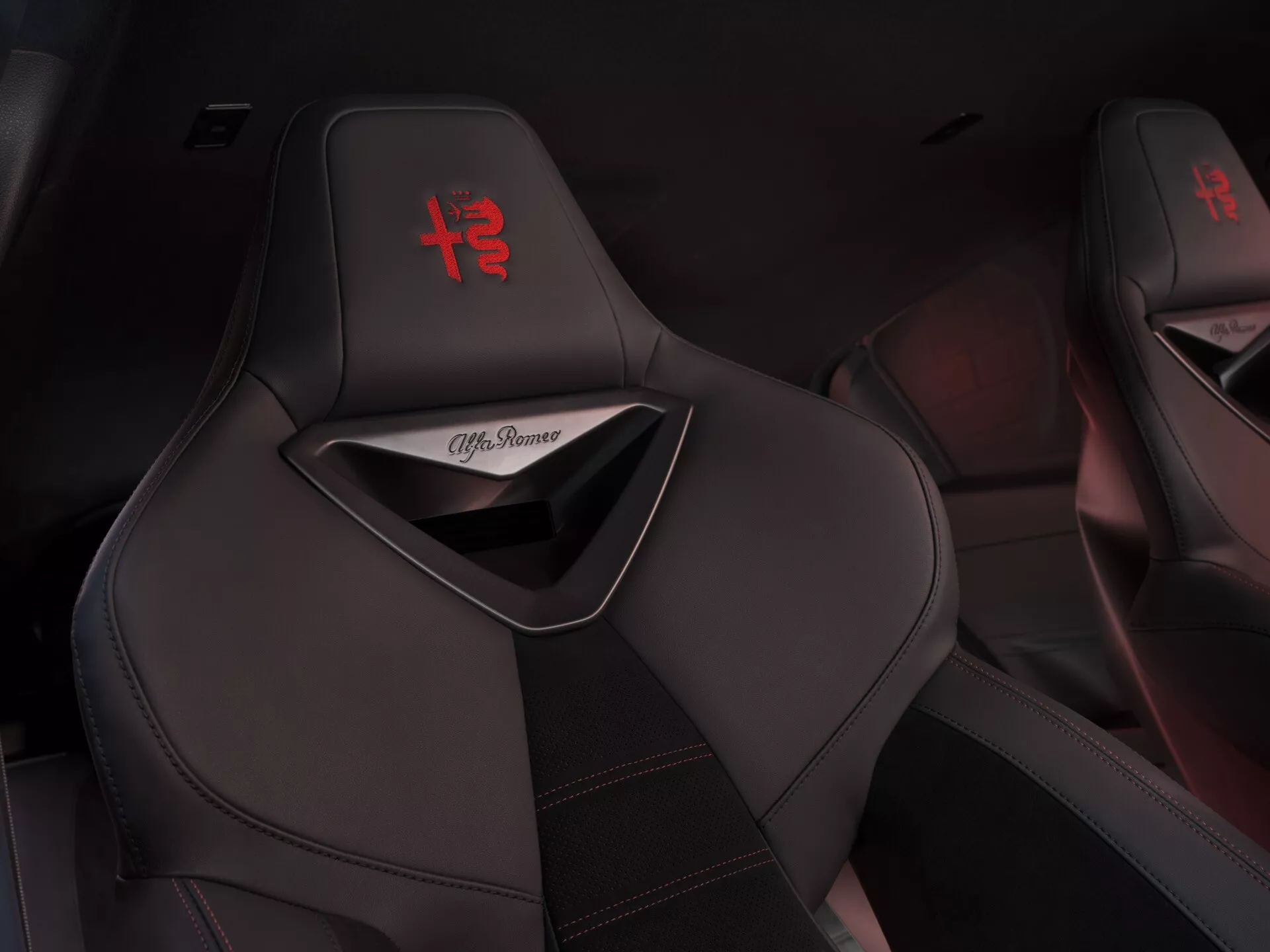 Zbliżenie na czarne sportowe fotele Alfa Romeo Junior 2024 z czerwonym logo '+' wyszywanym na zagłówku i czerwonymi przeszyciami kontrastowymi, prezentujące luksusowy i sportowy design wnętrza