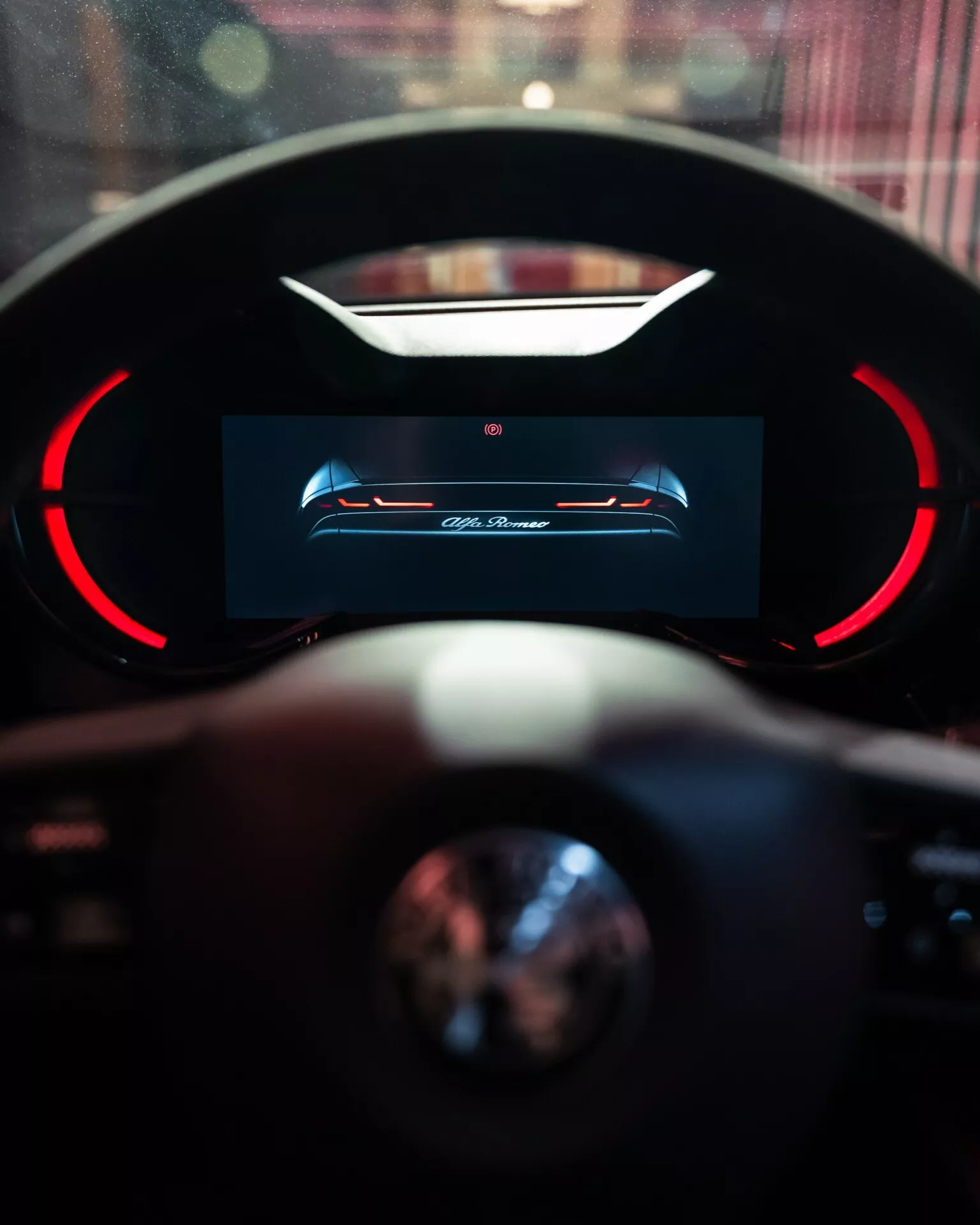 Widok wnętrza Alfa-Romeo Junior 2024 skupiający się na cyfrowym wyświetlaczu deski rozdzielczej kierowcy o wysokiej rozdzielczości z czerwonymi akcentami i częściowym widokiem kierownicy z rozmytym tłem wskazującym na luksusowe otoczenie