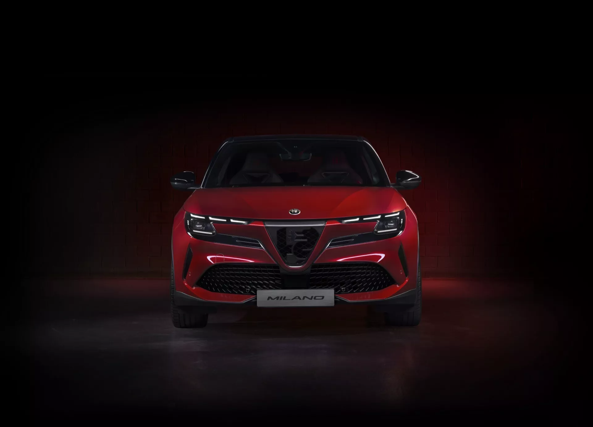 Przedni widok Alfa-Romeo Junior 2024 (966) w kolorze czerwonym, oświetlony w studio na ciemnym tle z subtelnym połyskiem poniżej, prezentujący charakterystyczną kratkę z dużymi wlotami powietrza, smukłe wąskie reflektory LED połączone pasem świetlnym, i odznakę edycji Milano, podkreślając jego sportowe i agresywne cechy projektowe.