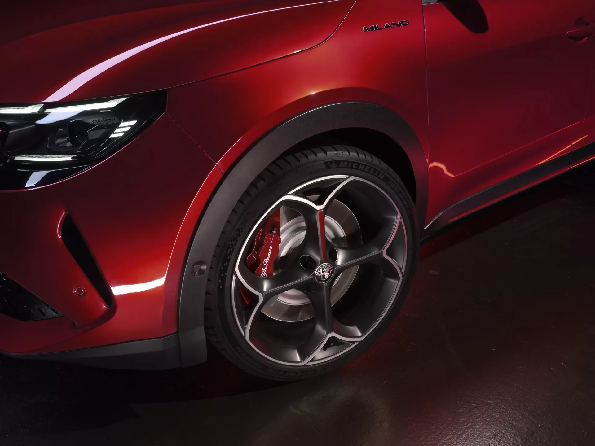 Zbliżenie na przednie lewe koło i błotnik Alfa Romeo Junior 2024, prezentujące wieloramienne felgi aluminiowe, błyszczący czerwony lakier i czerwony zacisk hamulcowy o wyglądzie premium i zorientowanym na osiągi