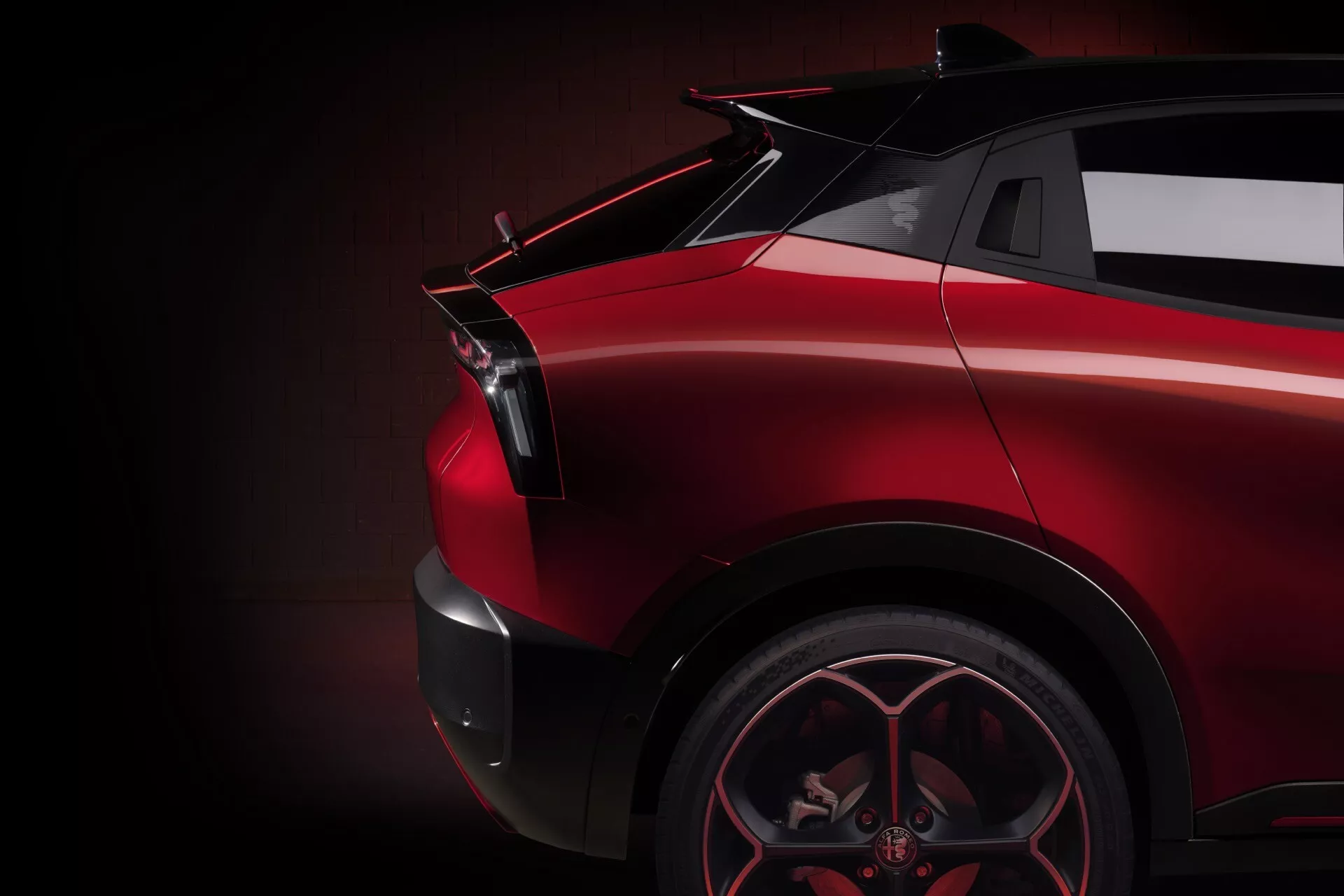 Zbliżenie boczne widoku Alfa Romeo Junior 2024 ukazujące tylną ćwiartkę po stronie pasażera ze szczególnym uwzględnieniem sportowego koła ze stopu, kątowego błotnika tylnego, rzeźbionego tylnego światła LED, lśniącego czerwonego wykończenia i możliwego spojlera na tle ciemnego, subtelnie oświetlonego tła podkreślającego dynamiczne i luksusowe elementy stylistyczne pojazdu.