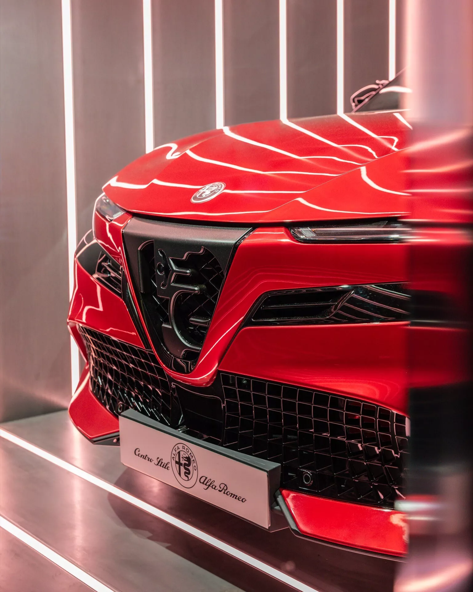 Zbliżenie na przednią lewą sekcję Alfa-Romeo Junior 2024 z żywym metalicznym czerwonym wykończeniem, ukazujące jej agresywny design, ostre kąty, rzeźbioną powierzchnię, wyraźny emblemat, dużą kątową grillę, eleganckie światła do jazdy dziennej i potencjalne wysokowydajne wloty powietrza, na prostym tle wnętrza, prawdopodobnie salonu.