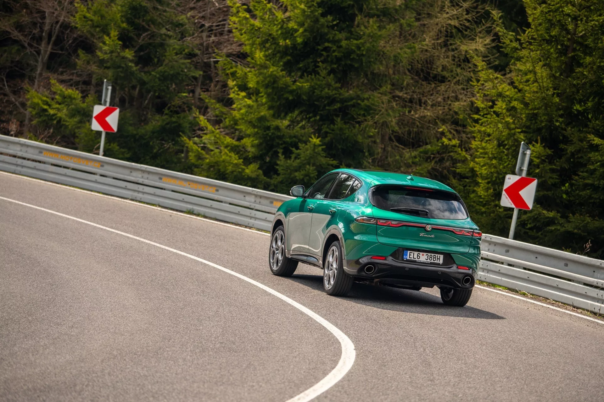 Alfa Romeo Tonale 2022 uchwycony z tylnego kąta trzech czwartych na zakrzywionej drodze, podkreślając jego sportowy design typu hatchback, błyszczące wykończenie i funkcje aerodynamiczne, w tym tylny spoiler, felgi aluminiowe i tylnie światła LED.