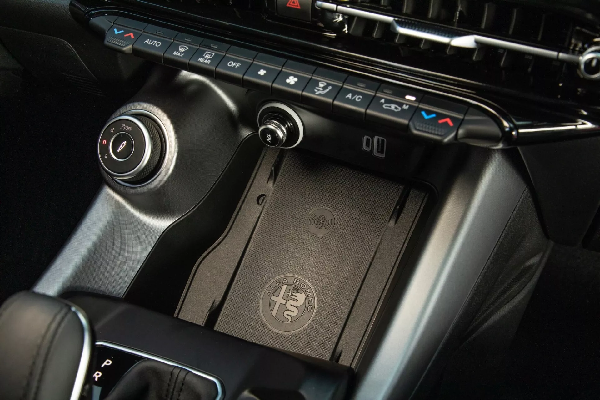 Zbliżenie na konsolę środkową Alfa Romeo Tonale 2022, prezentujące panel sterowania klimatyzacją, bezprzewodową stację ładującą, dźwignię zmiany biegów i pokrętło sterujące systemem rozrywki z wykończeniem premium i nowoczesnym designem.