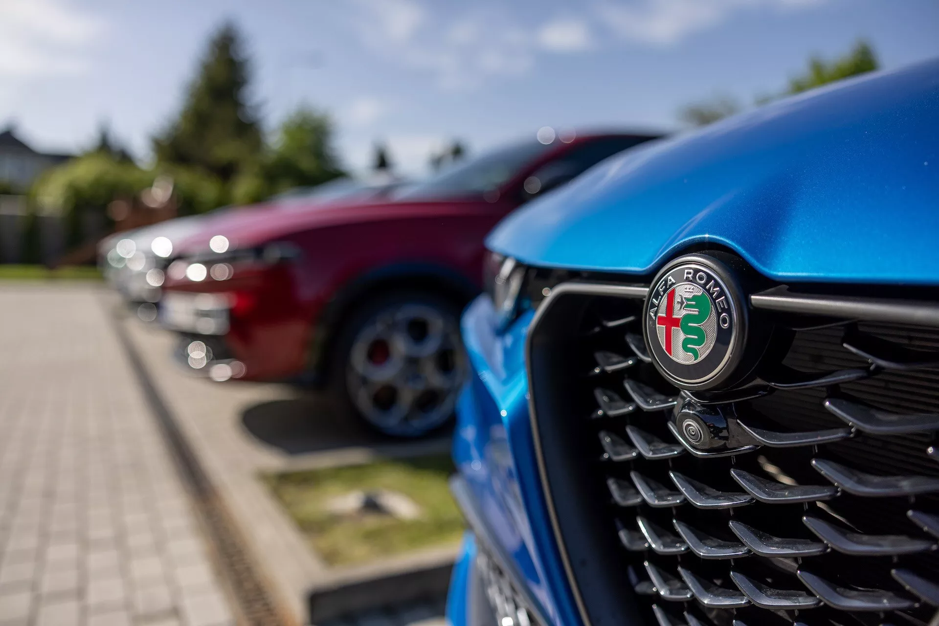 Zbliżenie na przednią kratę niebieskiego Alfa Romeo Tonale 2022 z naciskiem na emblemat, przedstawiające pionowe listwy i rozmyte tło innych pojazdów