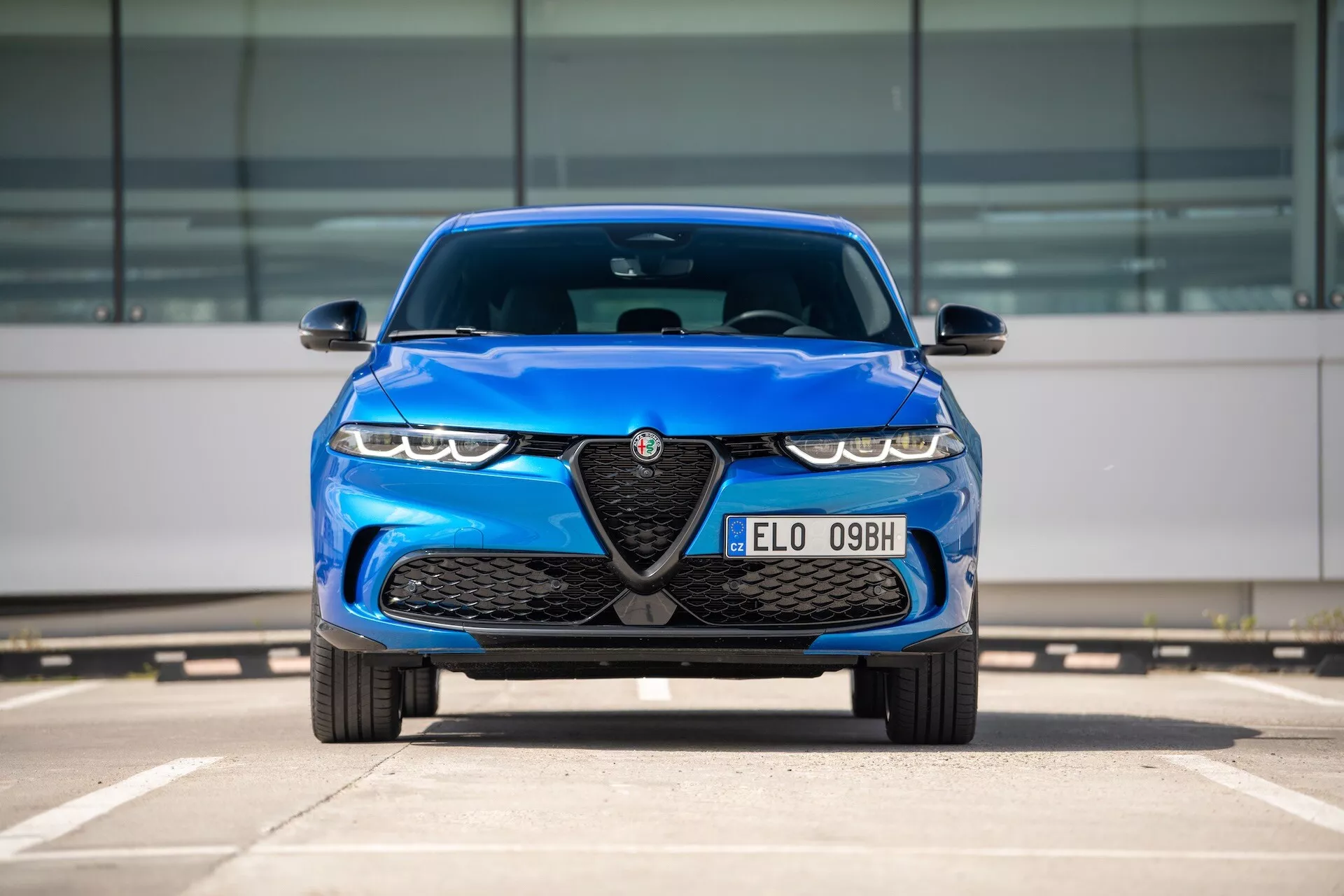 Niebieskie Alfa Romeo Tonale 2022, widok z przodu na parkingu prezentujący grill z emblematem, reflektory LED i cechy aerodynamiczne