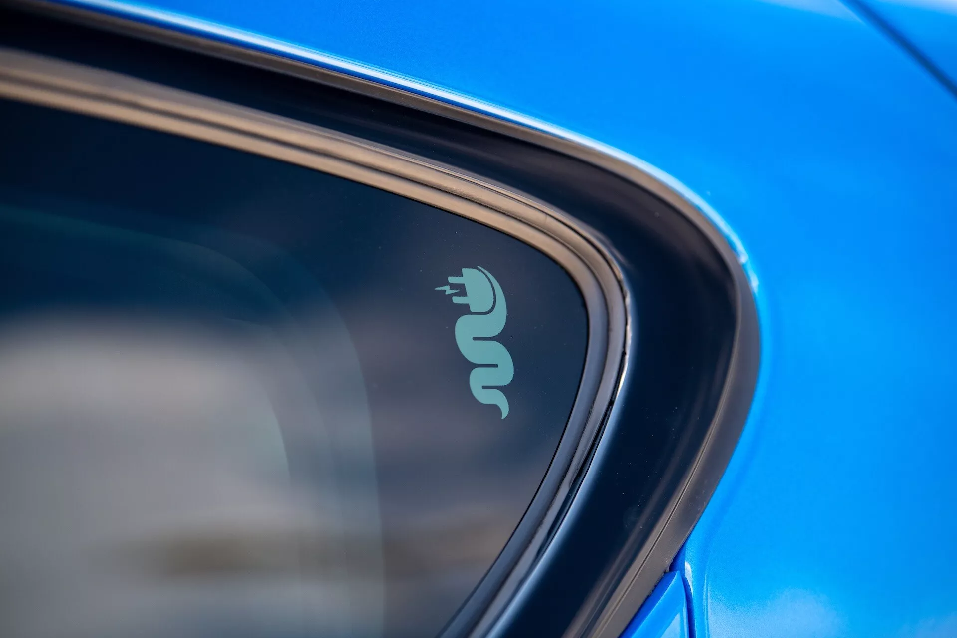 Szczegółowe zbliżenie na odznakę technologiczną Alfa Romeo Tonale 2022 wskazującą na elektryczną lub hybrydową zdolność w pobliżu okna samochodu, podkreśloną przez odblaskowy niebieski lakier i elegancki design w naturalnym świetle dzienym