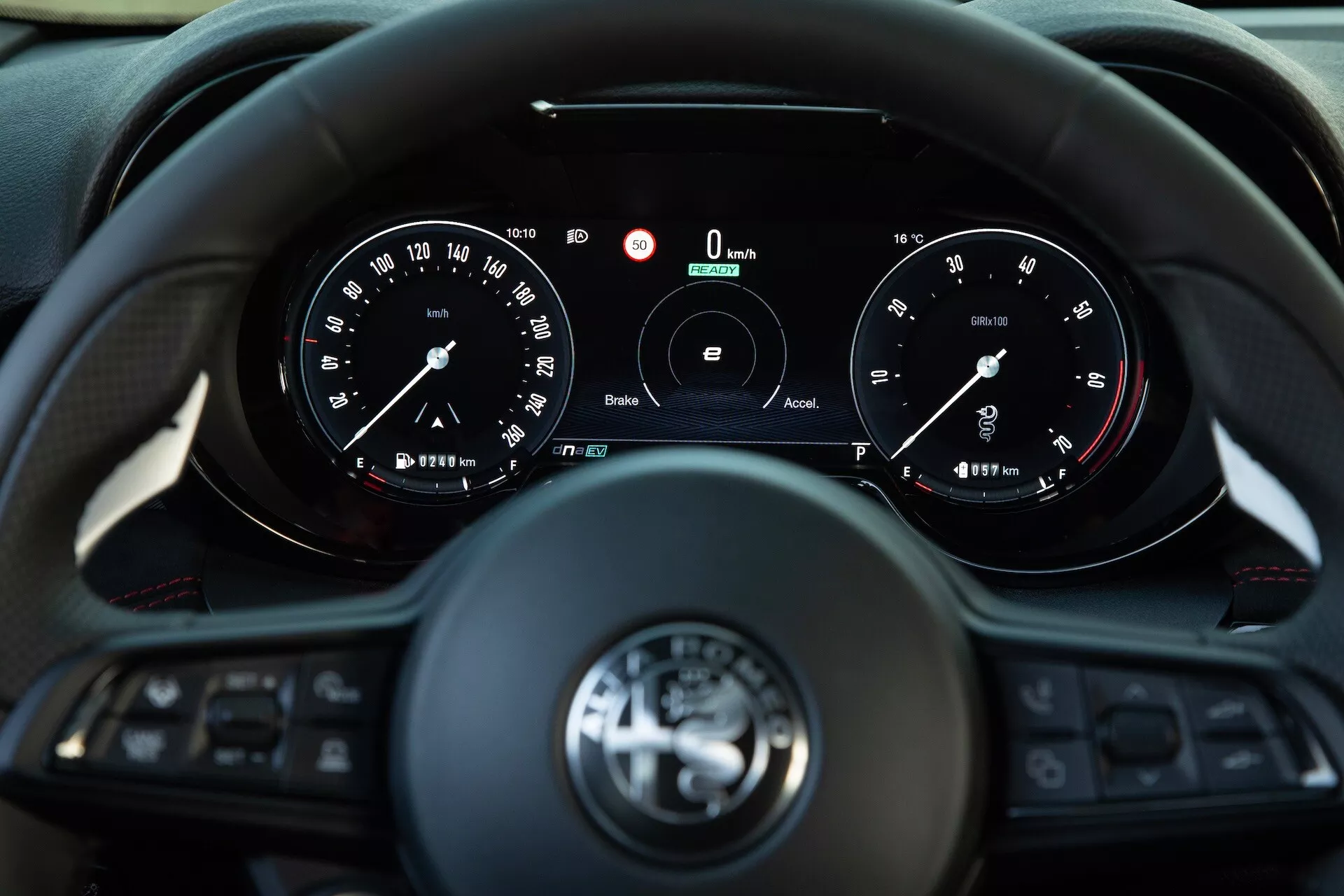 Wnętrze Alfa Romeo Tonale 2022 pokazujące widok kierowcy na wielofunkcyjną kierownicę o trzech ramionach i zestaw wskaźników z analogowymi tarczami i centralnym wyświetlaczem cyfrowym wskazującym prędkość i obroty na poziomie zero, zawierające technologie łączności i asysty kierowcy