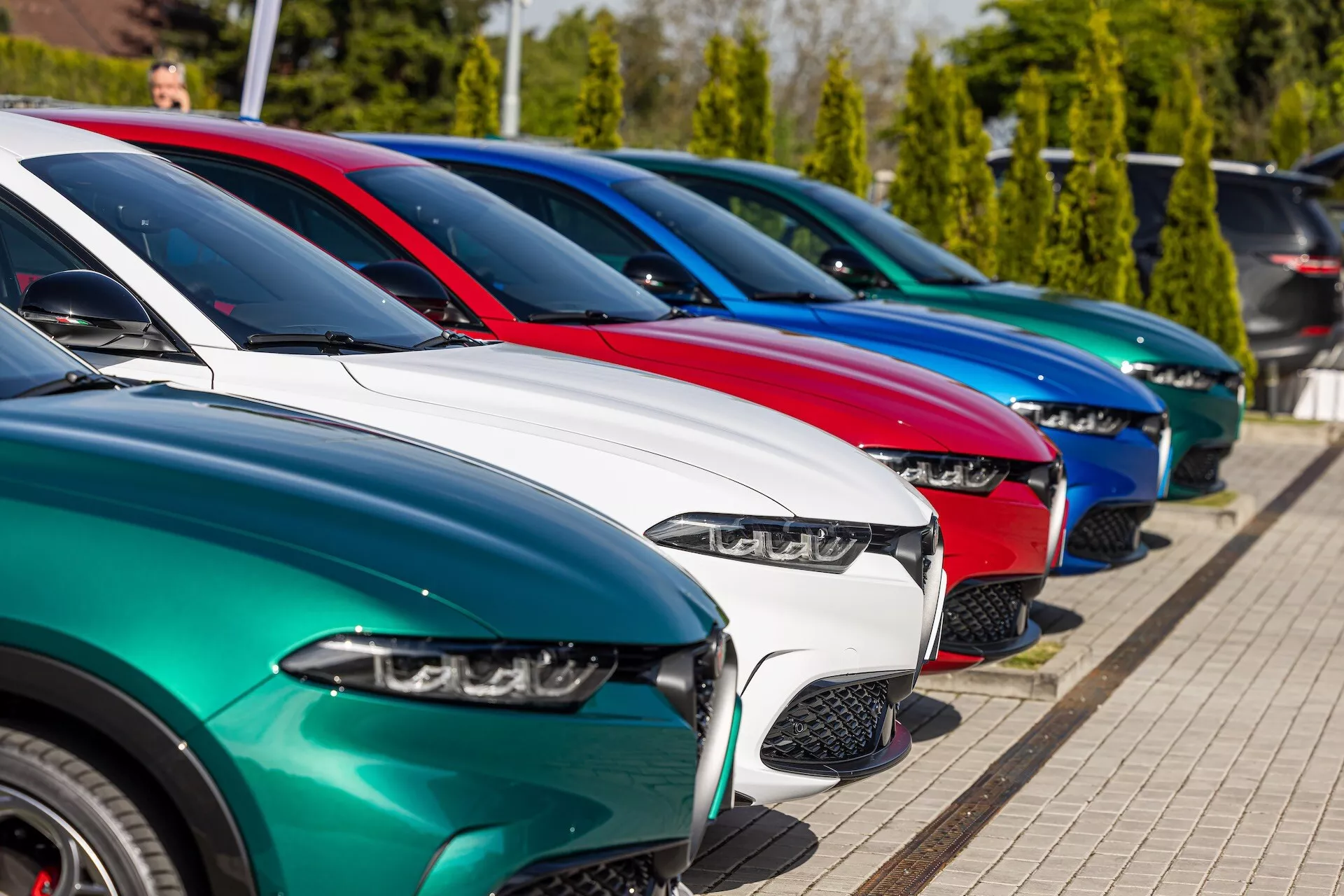 Rząd modeli Alfa Romeo Tonale 2022 zaparkowanych skośnie prezentujących widok z przodu z perspektywy trzech czwartych, podkreślających reflektory LED, zróżnicowane wzory grilla, lśniące wykończenia lakieru i koła ze stopów lekkich na tle jasnego słońca i zieleni
