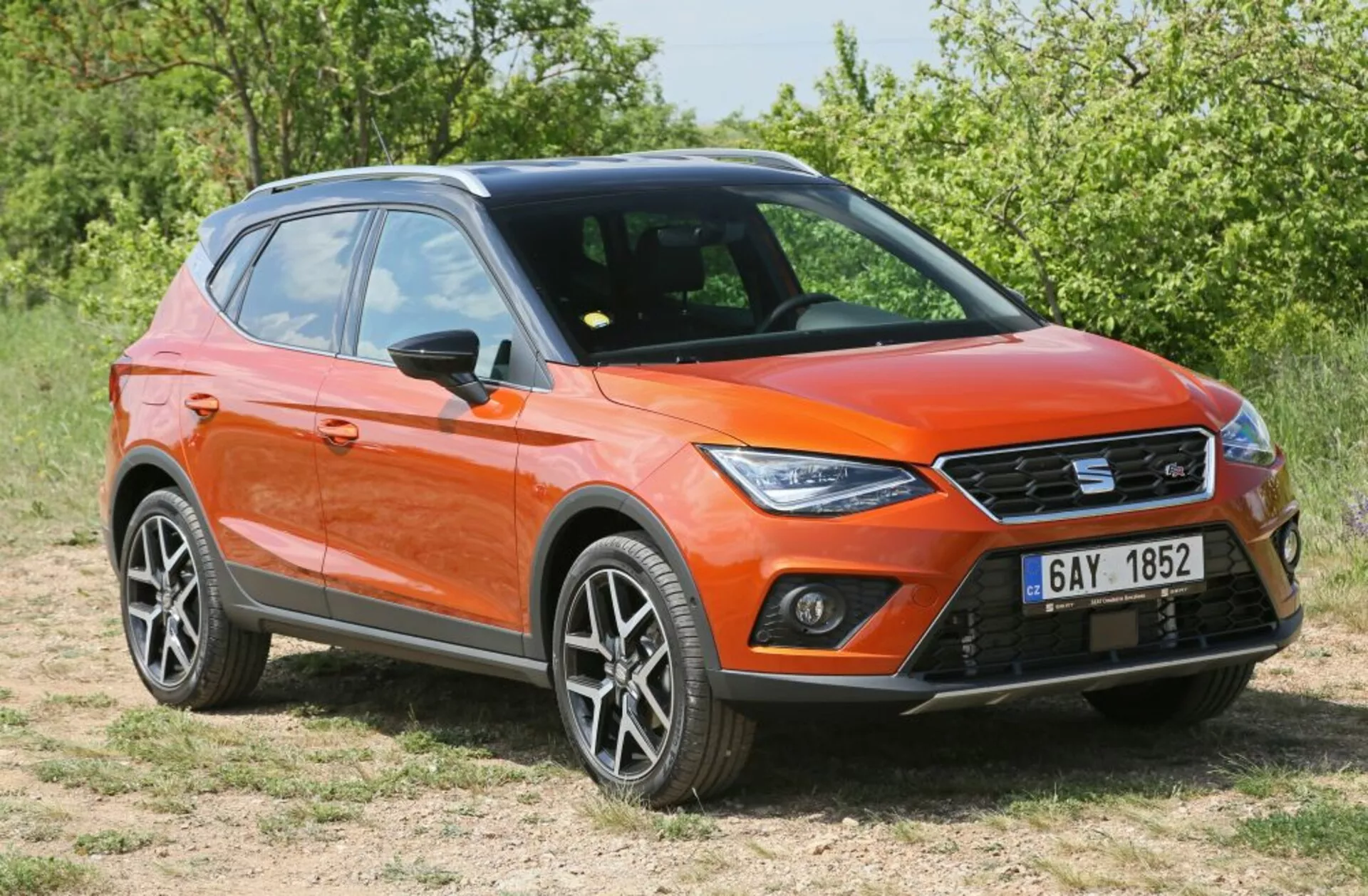 Pomarańczowy crossover/SUV zaparkowany na trawie w widoku trzy czwarte od przodu pokazujący LED-owe światła dzienne, nowoczesny ostry design z felgami aluminiowymi i relingami dachowymi w świetle słonecznym