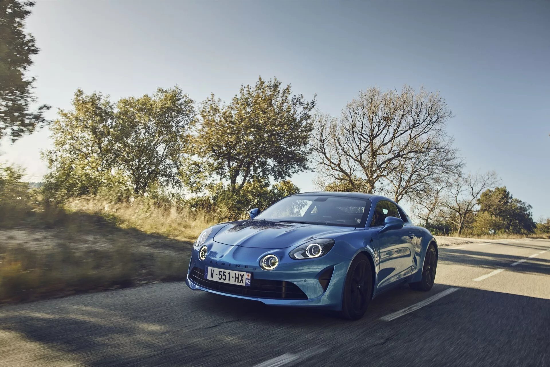 Dynamiczny trzy czwarte widok z przodu na niebieskie sportowe coupé Alpine A110 z 2017 roku w ruchu na wiejskiej drodze, podkreślający aerodynamiczny design, nowoczesne reflektory i sportową konfigurację kół