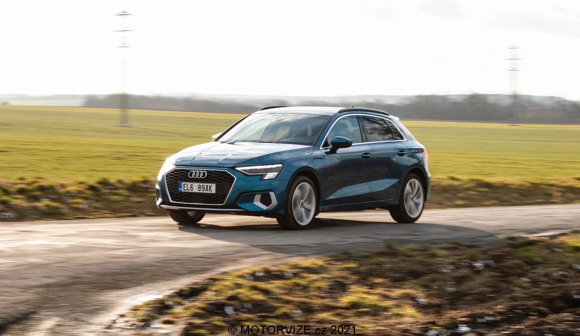 Dynamiczne ujęcie nowoczesnego hatchbacka, prawdopodobnie Audi A3 Sportback 2020, uchwyconego w ruchu. Obraz jest zrobiony pod lekko niskim kątem, podkreślając wysokość samochodu i tworząc dynamiczne wrażenie. Pokazuje widok z przodu pod kątem trzech czwartych, z samochodem poruszającym się od prawej do lewej. Charakterystyczne cechy projektowe obejmują eleganckie reflektory LED, dużą przednią kratkę, duże felgi aluminiowe, opływowe lusterka boczne z potencjalnymi wbudowanymi kierunkowskazami i metaliczne wykończenie lakierem z liniami konturowymi. Tło jest rozmyte, co zwiększa poczucie prędkości, podczas gdy sam samochód pozostaje w ostrym skupieniu, podkreślając jego nowoczesny design i premium estetykę.