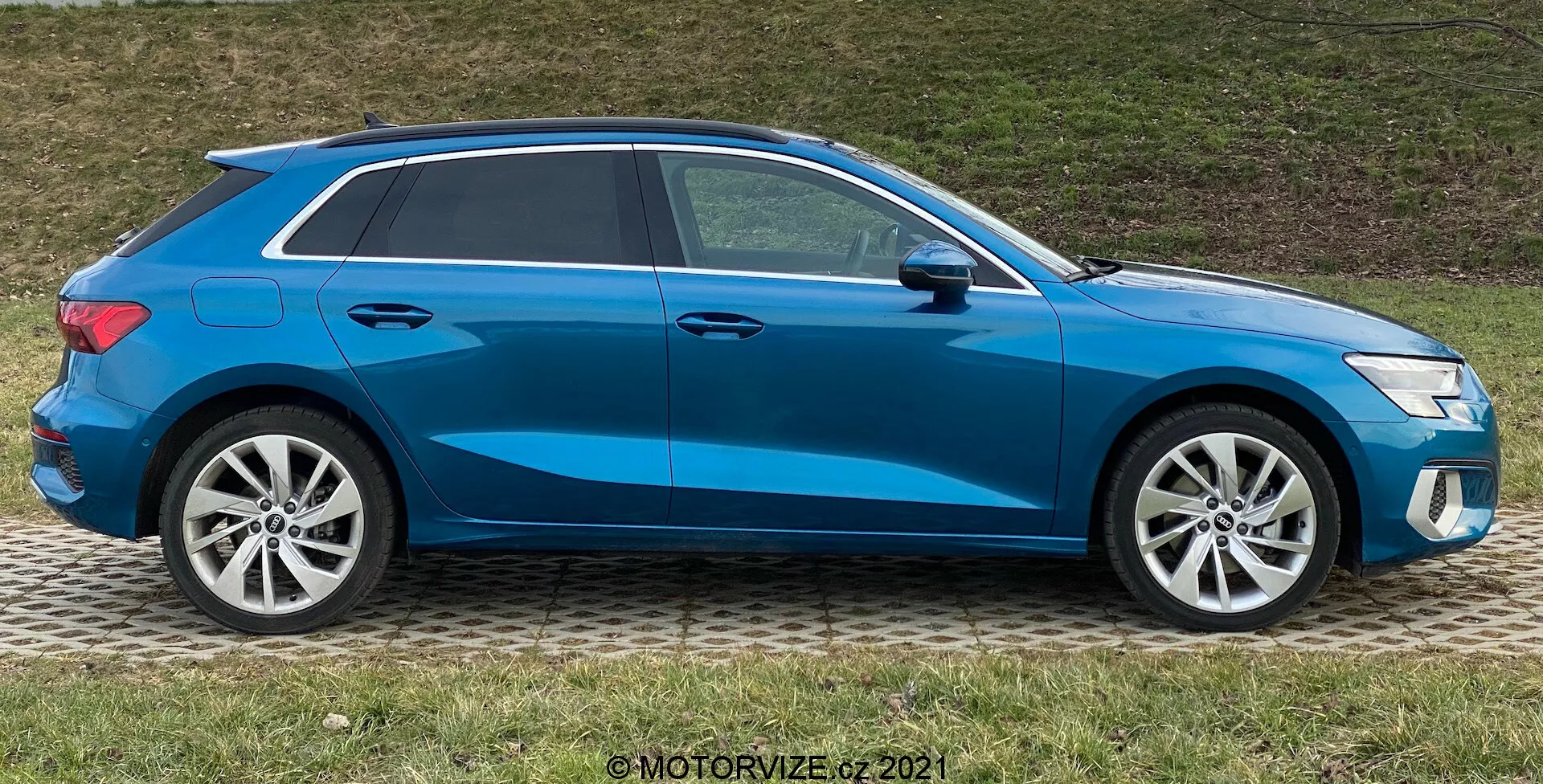 Widok boczny niebieskiego Audi A3 Sportback z 2020 roku, prezentujący nowoczesny hatchback z sportowym opadającym dachem, felgami aluminiowymi, zamkniętymi oknami i bocznymi lusterkami w kolorze nadwozia z wbudowanymi kierunkowskazami, zaparkowany na trawie obok chodnika.