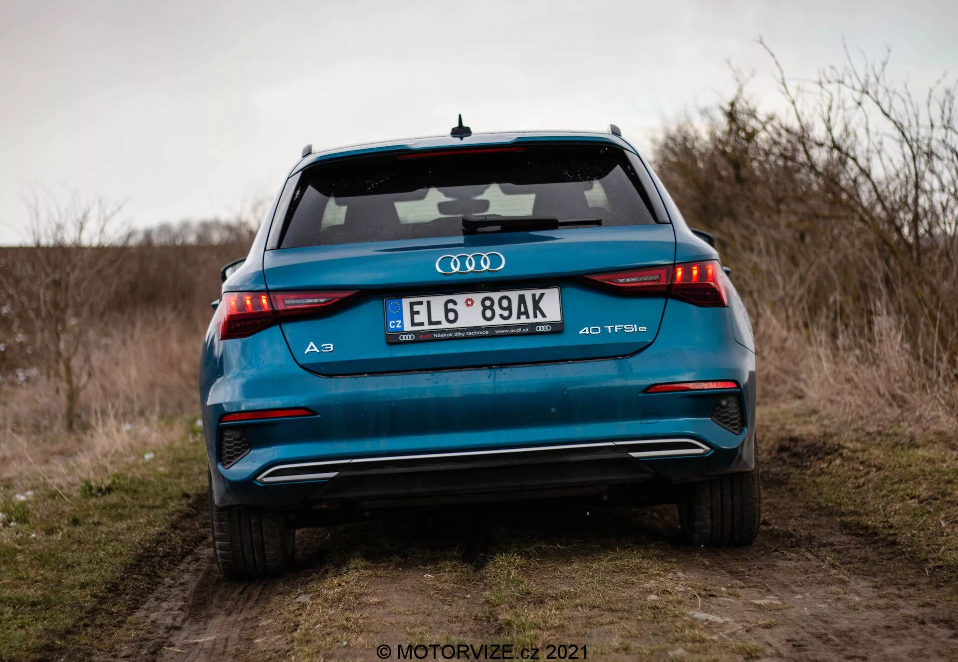 Szczegółowy widok tylnej części jaskrawo niebieskiego Audi A3 Sportback 40 TFSI z 2020 roku, podkreślający jego zaawansowane cechy projektowe, w tym poziome tylne światła LED, podwójne końcówki wydechu, emblem turbodoładowanego silnika TFSI, zintegrowany spoiler i antenę płetwową rekina, sfotografowany pod niskim kątem na polnej drodze z otwartym polem i zachmurzonym niebem w tle.