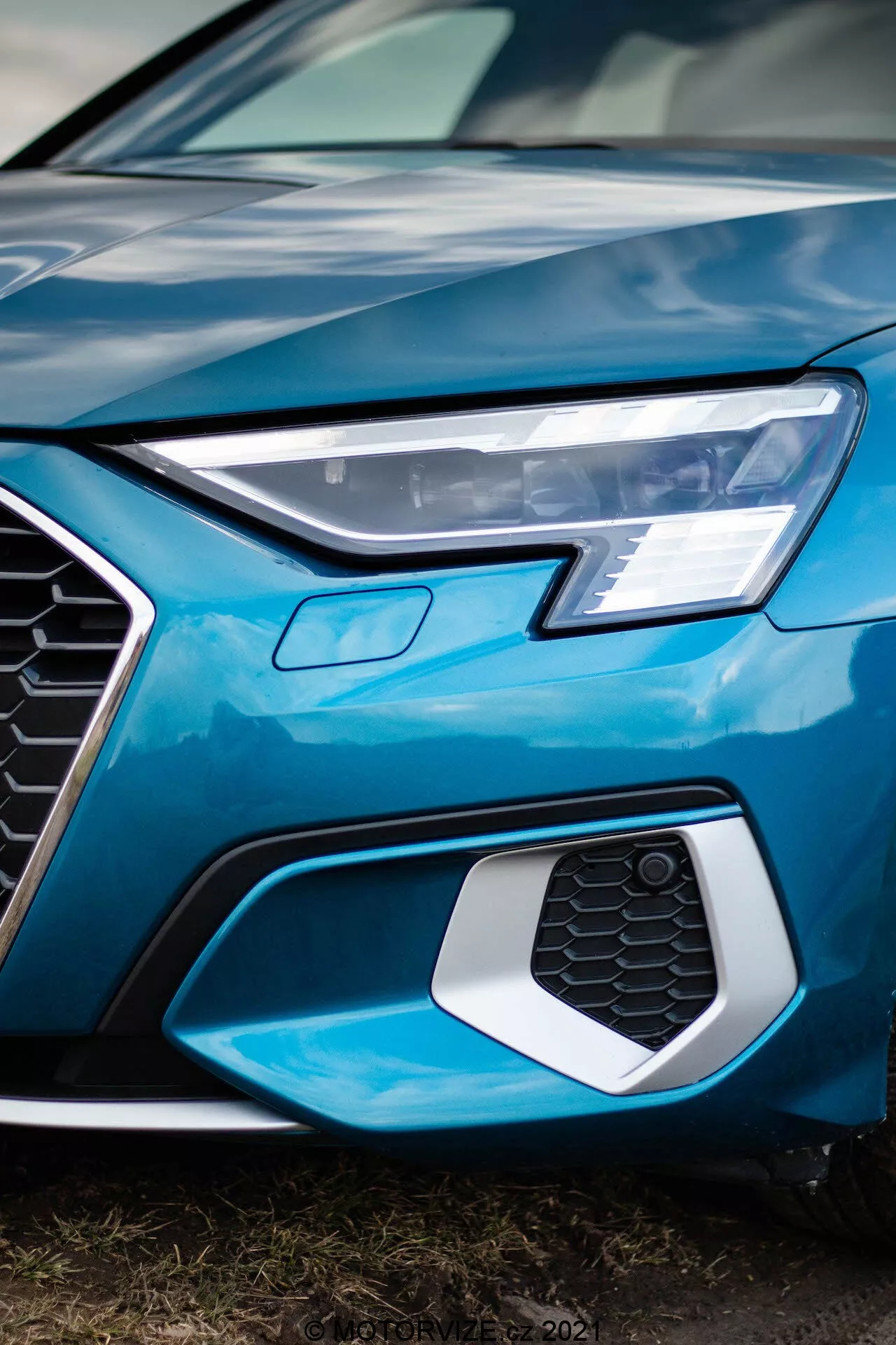 Zbliżenie, niskokątny kadr przedniej lewej części Audi A3 Sportback 2020, ukazujący ostre reflektory LED, sześciokątną kratkę w kształcie plastra miodu mogącą mieścić czujniki lub emblematy, oraz część błyszczącej jasno- niebieskiej maski w, podkreślając nowoczesny, wyszukany design i zaawansowaną technologię samochodową.