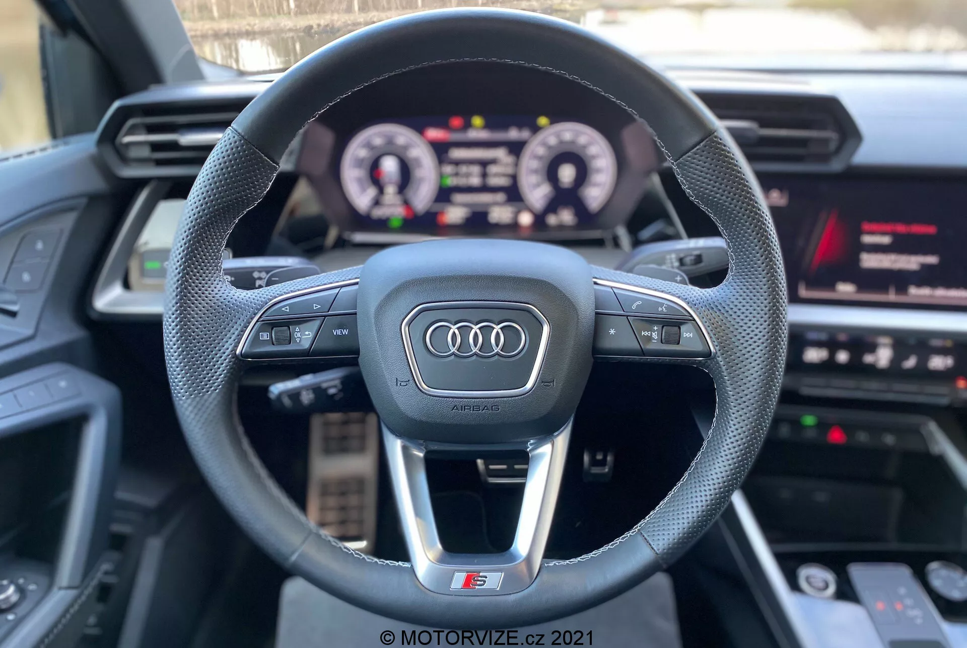 Perspektywa pierwszoosobowa z fotela kierowcy w Audi A3 2020 (A3 Sportback) ukazująca sportową kierownicę o trzech ramionach z logo Audi, wielofunkcyjnymi przyciskami kontrolnymi, podświetloną cyfrową deską rozdzielczą oraz częścią systemu informacyjno-rozrywkowego z multimediami, nawigacją i ustawieniami pojazdu. Funkcje podkreślają interaktywną technologię i luksusowe komponenty z naciskiem na wrażenia z jazdy.