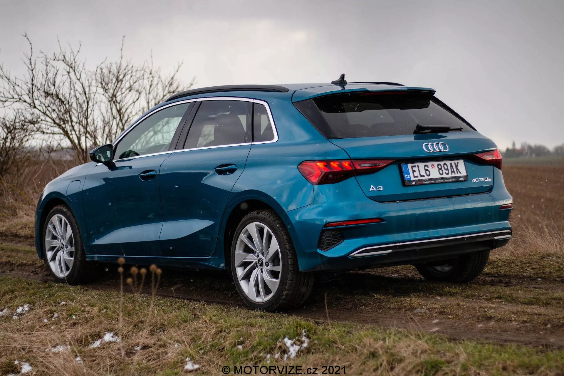 Niebieskie Audi A3 2020 (Sportback) sfotografowane z tyłu pod kątem trzech czwartych na otwartym polu pod pochmurnym niebem. Obraz uwidacznia prawą stronę samochodu, tylne światła LED, podwójny układ wydechowy, wieloramienne felgi aluminiowe i tylny spojler. Pojazd jest zaparkowany na nieutwardzonej powierzchni z kępami trawy i odrobiną śniegu lub szronu, co sugeruje zimne warunki pogodowe. Ogólna atmosfera jest ponura, z naciskiem na sportowy i ekskluzywny wygląd Audi A3.