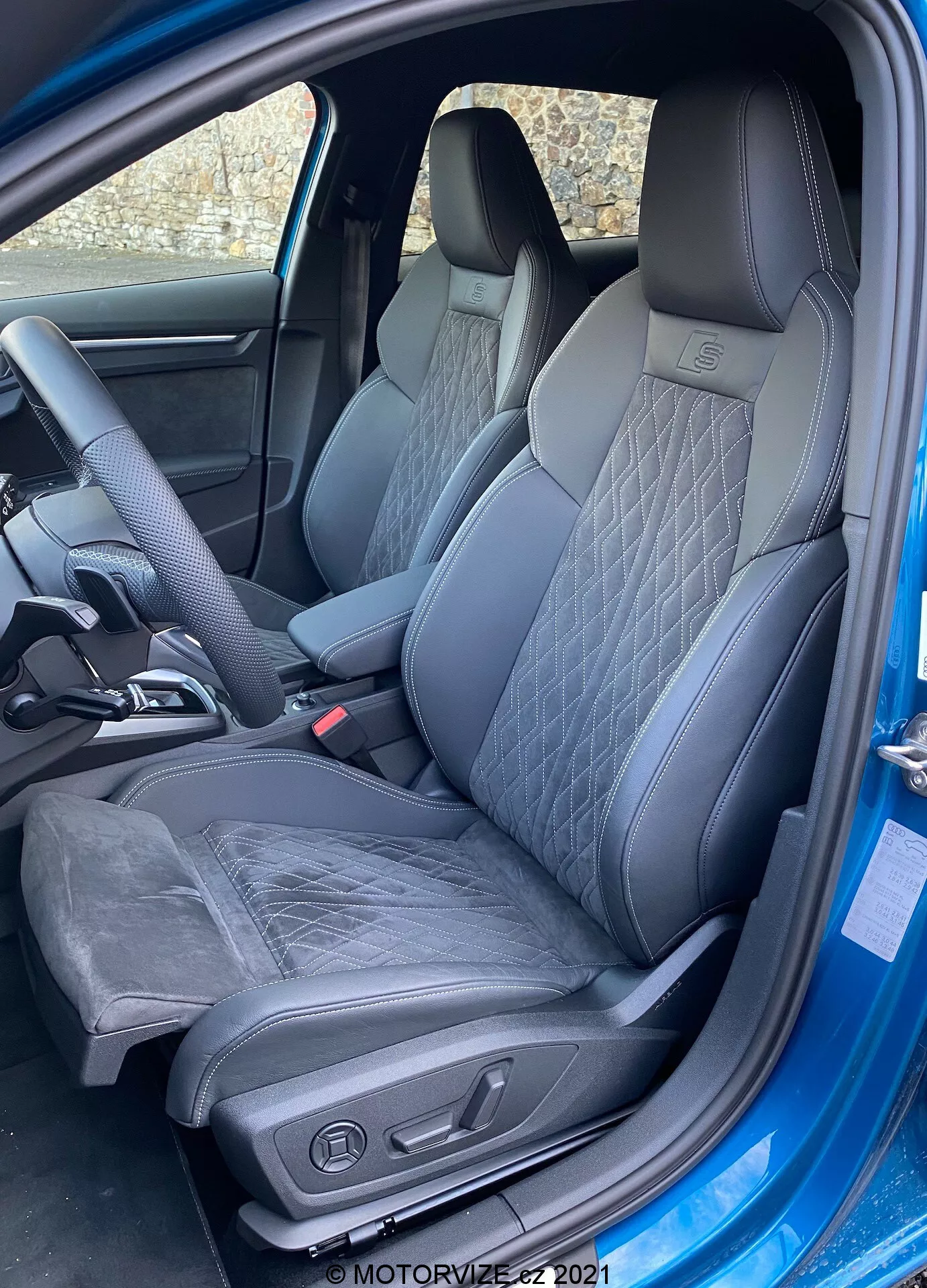 Zewnętrzne ujęcie w średnim kącie wnętrza Audi A3 Sportback 2020, z naciskiem na fotel kierowcy. Drzwi są całkowicie otwarte, odsłaniając sportowy, nowoczesny fotel wykonany ze skóry i być może Alcantary. Fotel ma pikowane wzory i wyraźne boczne wzmocnienia zapewniające wsparcie boczne. Widoczne są elektryczne sterowniki regulacji fotela, sugerujące regulację wysokości, nachylenia i podparcia lędźwiowego. Wnętrze jest monochromatyczne z ciemnymi tonami, co przyczynia się do ekskluzywnego wyglądu. Widoczna jest również część ergonomicznej kierownicy z miejscem na kciuk, co wskazuje na sportową kierownicę.