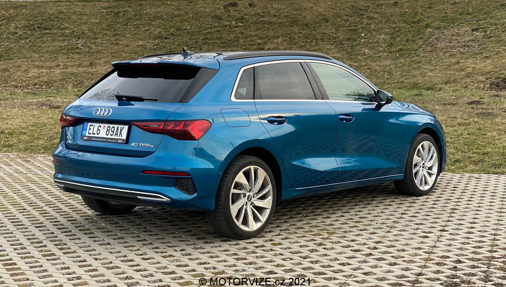 Obraz przedstawia niebieskie Audi A3 2020 Sportback z trzy czwarte tylnego widoku, podkreślając prawe tylne partie. Kluczowe szczegóły obejmują wieloramienne felgi aluminiowe ze srebrnym wykończeniem, częściowo widoczne diodowe światła tylne, smukłą aerodynamikę z wyraźną linią ramion, oznakowanie tylne wskazujące model i być może poziom wyposażenia, ciemniejsze szyby tylne zapewniające prywatność, mały spoiler dachowy, relingi dachowe dla uniwersalności i podwójne końcówki wydechów w dolnym tylnym zderzaku. Samochód jest zaparkowany na wzorzystej powierzchni z trawiastym tłem, co sugeruje otoczenie nie zbyt miejskie.
