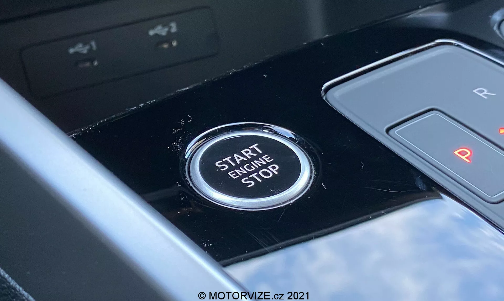 Zbliżenie na przycisk start/stop silnika w Audi A3 Sportback 2020 umieszczony w centralnej konsoli. Przycisk wyraźnie pokazuje tekst 'START ENGINE STOP' wielkimi literami, wskazując jego funkcję zapalania lub gaszenia silnika. Widok z perspektywy kierowcy, błyszczący czarny panel i częściowo widoczna dźwignia zmiany biegów oznaczona 'R' i 'P' sugerują automatyczną skrzynię biegów. Płytka głębia ostrości podkreśla wyrazistość przycisku na tle lekko rozmytym, wskazując na nowoczesne wnętrze pojazdu z zaawansowanymi funkcjami technologicznymi.