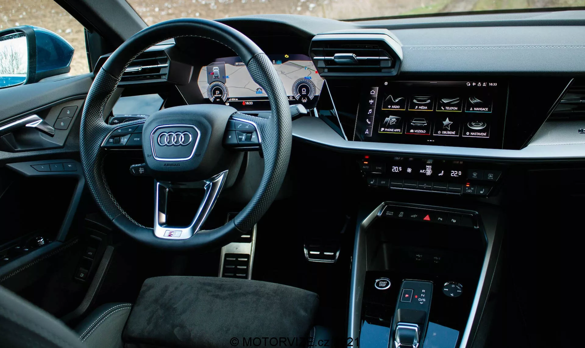 Widok wnętrza Audi A3 Sportback 2020 z lekko zdecentralizowanego lewego kąta, skupiający się na stronie kierowcy. Wyraźny w centrum zdjęcia znajduje się kierownica z logo czterech pierścieni Audi, wielofunkcyjnymi przyciskami i widocznymi łopatkami zmiany biegów. Za kierownicą znajduje się cyfrowa tablica rozdzielcza, czyli Audi Virtual Cockpit, pokazująca prędkościomierz, obrotomierz i mapę nawigacyjną. Konsola środkowa obejmuje poziomy ekran dotykowy dla systemu multimedialnego, interfejs sterowania klimatyzacją, przycisk start-stop silnika, inny ekran dotykowy do sterowania klimatyzacją i minimalistyczny selektor biegów. Wnętrze cechuje się wysokiej jakości materiałami oraz technologicznie zintegrowanym designem o prestiżowym wyglądzie.