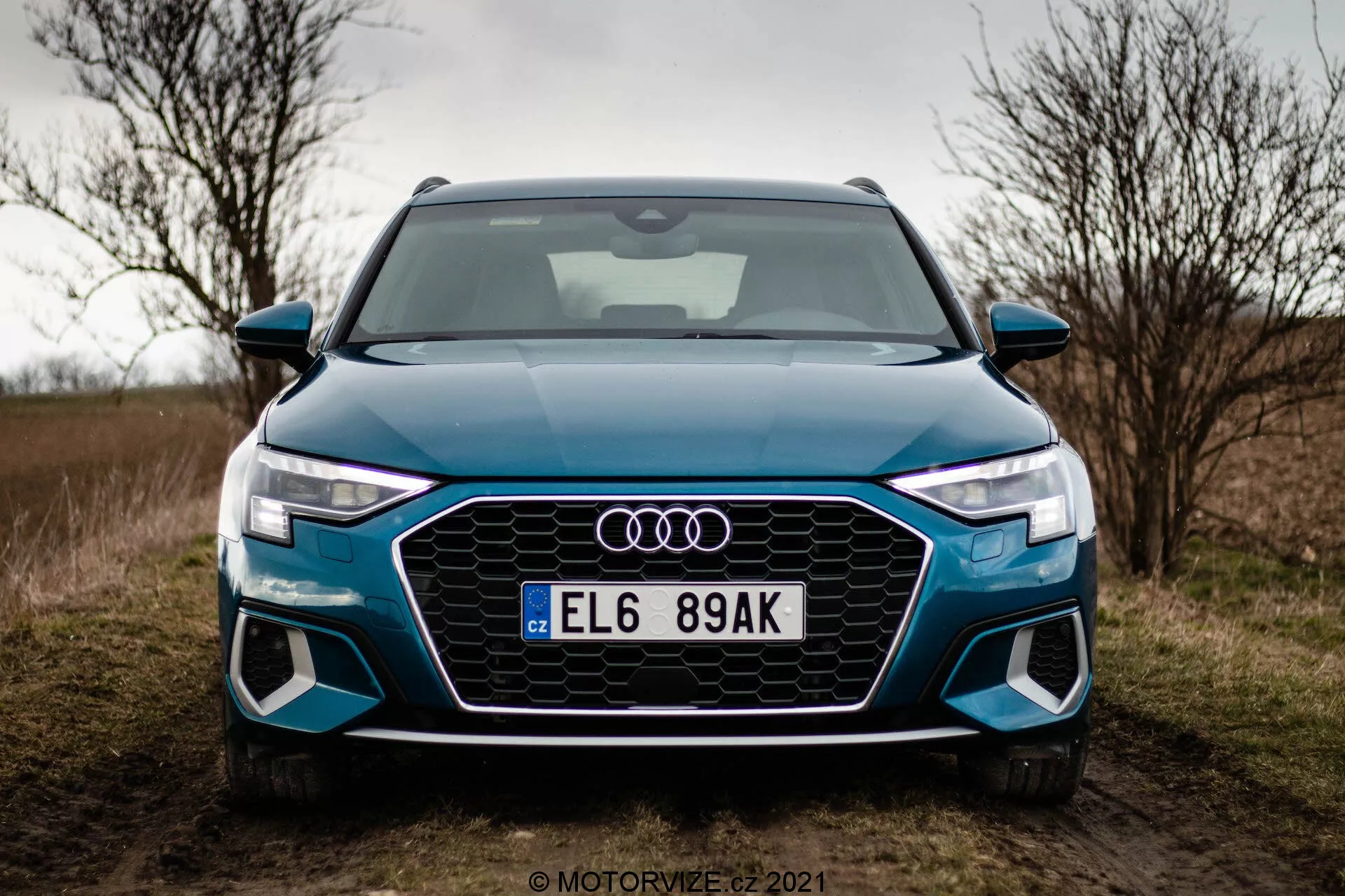 Widok z przodu Audi A3 Sportback z 2020 roku sfotografowany z niskiego kąta. Obraz koncentruje się na przedniej części samochodu, prezentując oktagonalną osłonę Singleframe z wstawkami w kształcie plastra miodu, ostre, kątowe reflektory LED z charakterystycznymi światłami do jazdy dziennej oraz duże wloty powietrza w rzeźbionym przednim zderzaku. Cztery pierścienie Audi są zamontowane centralnie na osłonie, z europejską tablicą rejestracyjną poniżej. Samochód, w jasnym odcieniu niebieskiego, jest zaparkowany przy trawiastej drodze z pochmurnym niebem w tle, sugerując jesienną lub zimową porę roku.