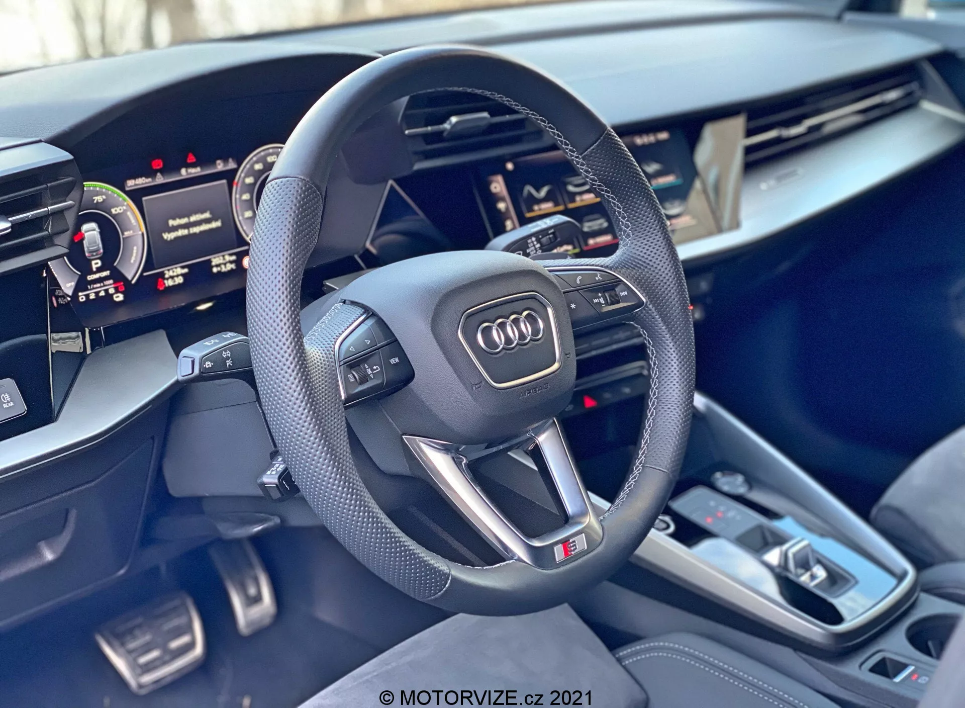 Wnętrze Audi A3 Sportback 2020, skupiające się na kierownicy i konsoli centralnej. Sportowa, trójramienna kierownica z płaskim dołem ma perforowane skórzane uchwyty, wielofunkcyjne przyciski, emblemat Audi i znaczek 'S'. Za kierownicą częściowo widoczny jest cyfrowy zestaw wskaźników, a z prawej strony częściowo widoczny jest dotykowy wyświetlacz Centralnego Interfejsu Multimedialnego (MMI). Konsola centralna zawiera dźwignię zmiany biegów automatycznej skrzyni biegów, wiele przycisków, touchpad, dwa uchwyty na kubki i małe schowki. Materiały wykończeniowe wnętrza cechują się premium akcentami szczotkowanego metalu i wysokiej jakości wykończeniami, podkreślając luksus i zaawansowaną technologię samochodu.