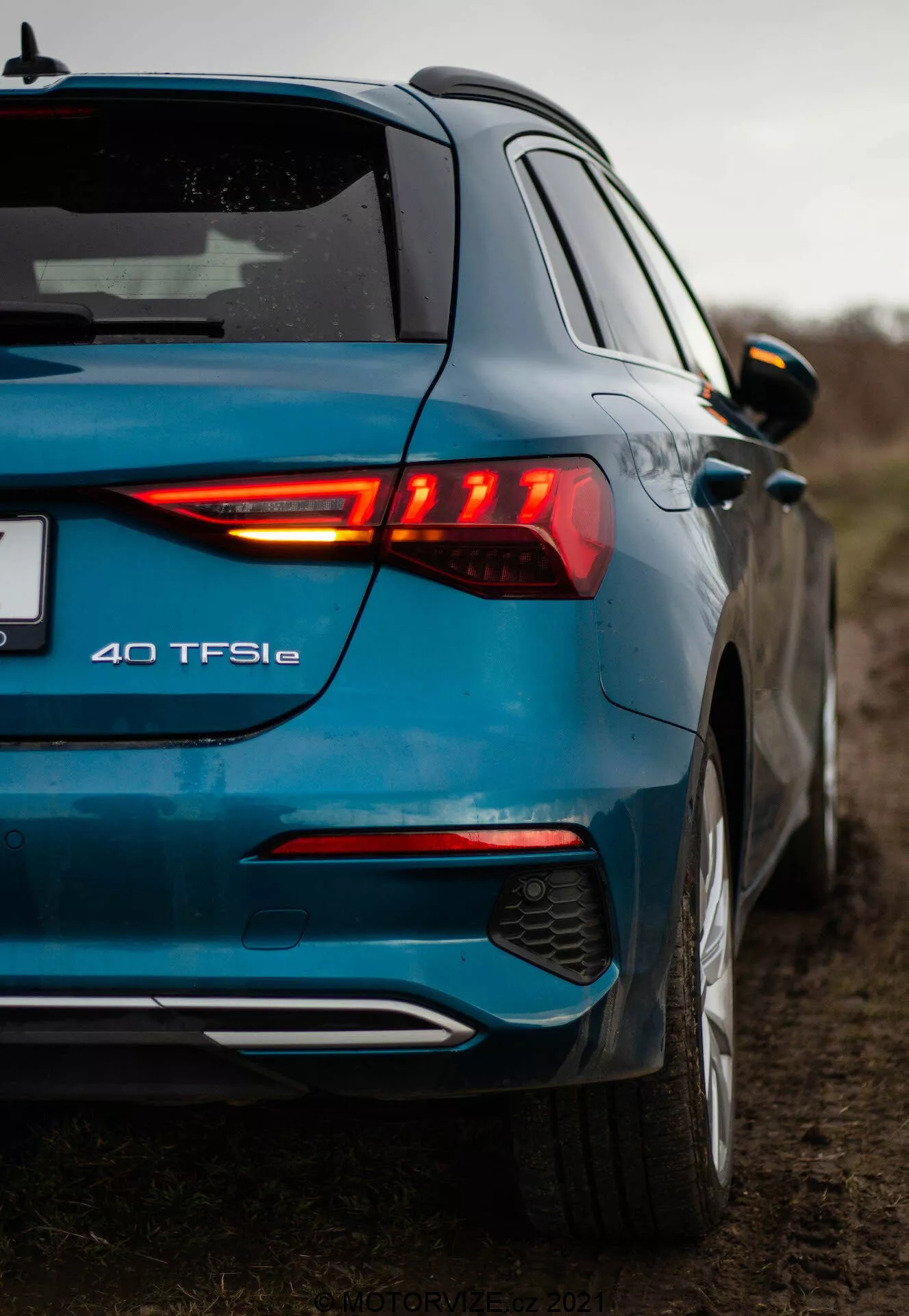 Zdjęcie niskiego kąta nowoczesnego niebieskiego Audi A3 2020 (A3 Sportback) sfotografowane od tyłu z trzech czwartych, podkreślające podświetlony tylny klaster LED, oznaczenie modelu 40 TFSIe, dwukolorowy tylny zderzak z zintegrowanym dyfuzorem i czujnikami parkowania, metaliczne wykończenie lakieru, spojler dachowy, lusterka boczne w kolorze nadwozia z zintegrowanymi kierunkowskazami oraz zarys przednich drzwi i bocznego profilu. Fotografia podkreśla elementy designu i oznakowanie samochodu w wiejskim środowisku z zachmurzonym niebem i małą głębią ostrości.