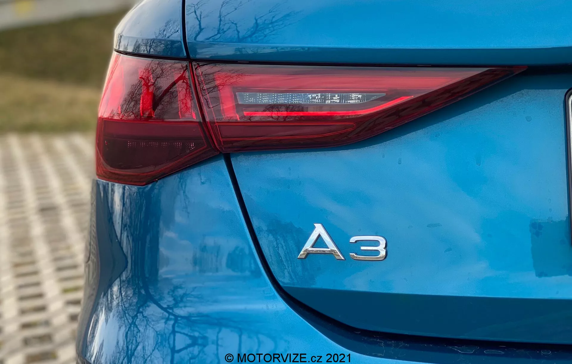 To zdjęcie przedstawia zbliżenie tylnego lewego narożnika niebieskiego Audi A3 Sportback 2020, wykonanego pod niskim kątem. Fotografia podkreśla elegancki design tylnej lampy, która ma ostrą, poziomą orientację rozciągającą się na bok pojazdu, oraz chromowane oznaczenie modelu 'A3' tuż poniżej. Elementy LED wewnątrz tylnej lampy podkreślają zaawansowaną technologię oświetleniową auta. Błyszczący lakier i ogólna czystość modelu ukazują wysokiej jakości detale samochodu, z lekko rozmytym tłem sugerującym otoczenie na świeżym powietrzu. Znak wodny '© MOTORVIZE.cz 2021' wskazuje na własność i pochodzenie zdjęcia.
