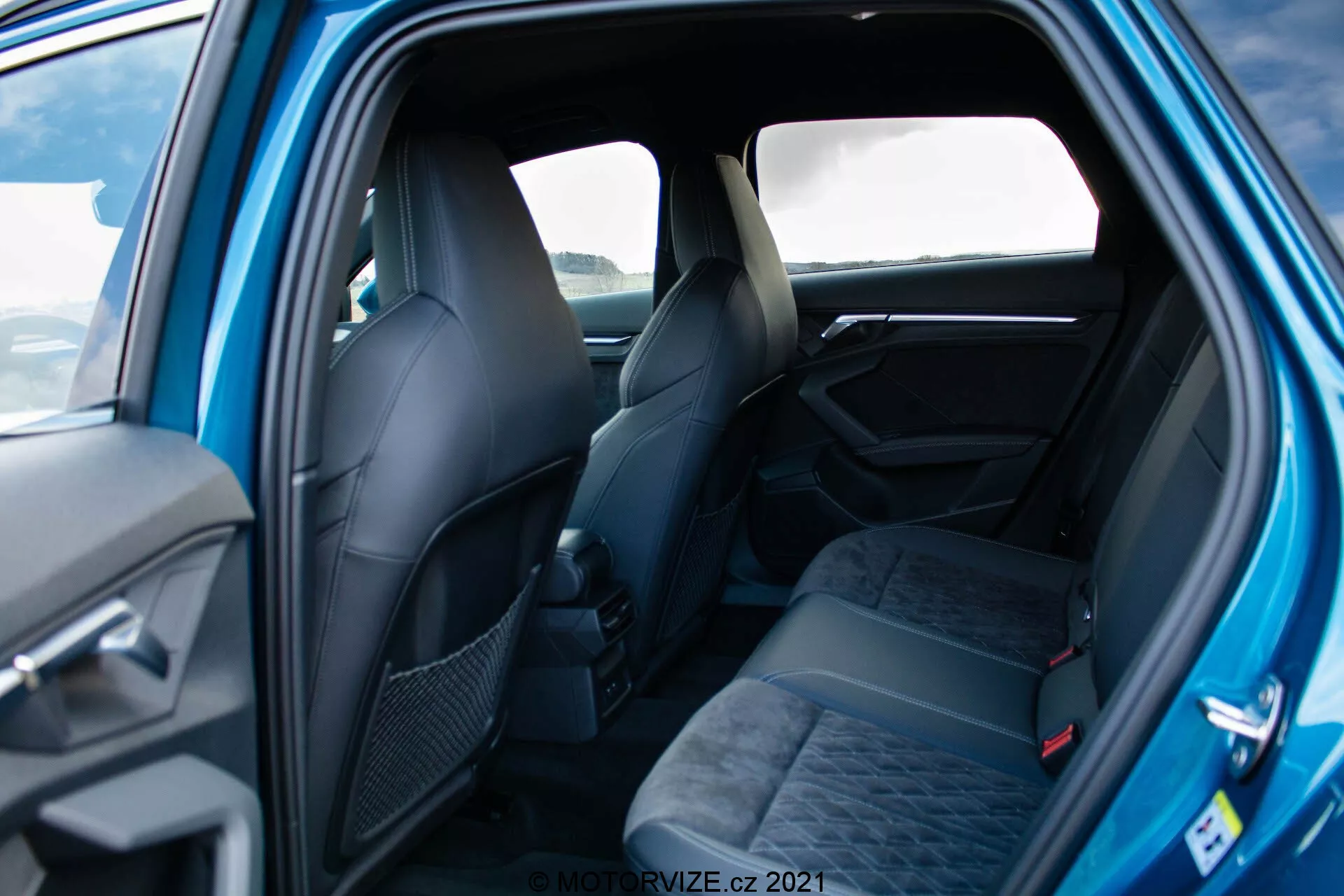 Zdjęcie wnętrza Audi A3 Sportback 2020 z perspektywy tylnego pasażera, ukazujące przestronne siedzenia dla trzech osób z zagłówkami i pasami bezpieczeństwa, tapicerowane ciemną tkaniną z wzorem. Częściowo widoczne są przednie siedzenia o ergonomicznym kształcie. Widoczne elementy panelu drzwiowego obejmują uchwyt i możliwe przyciski sterujące. Dobrze oświetlone zdjęcie ukazuje elegancki, wysokiej jakości wygląd z użyciem charakterystycznych materiałów i detali.