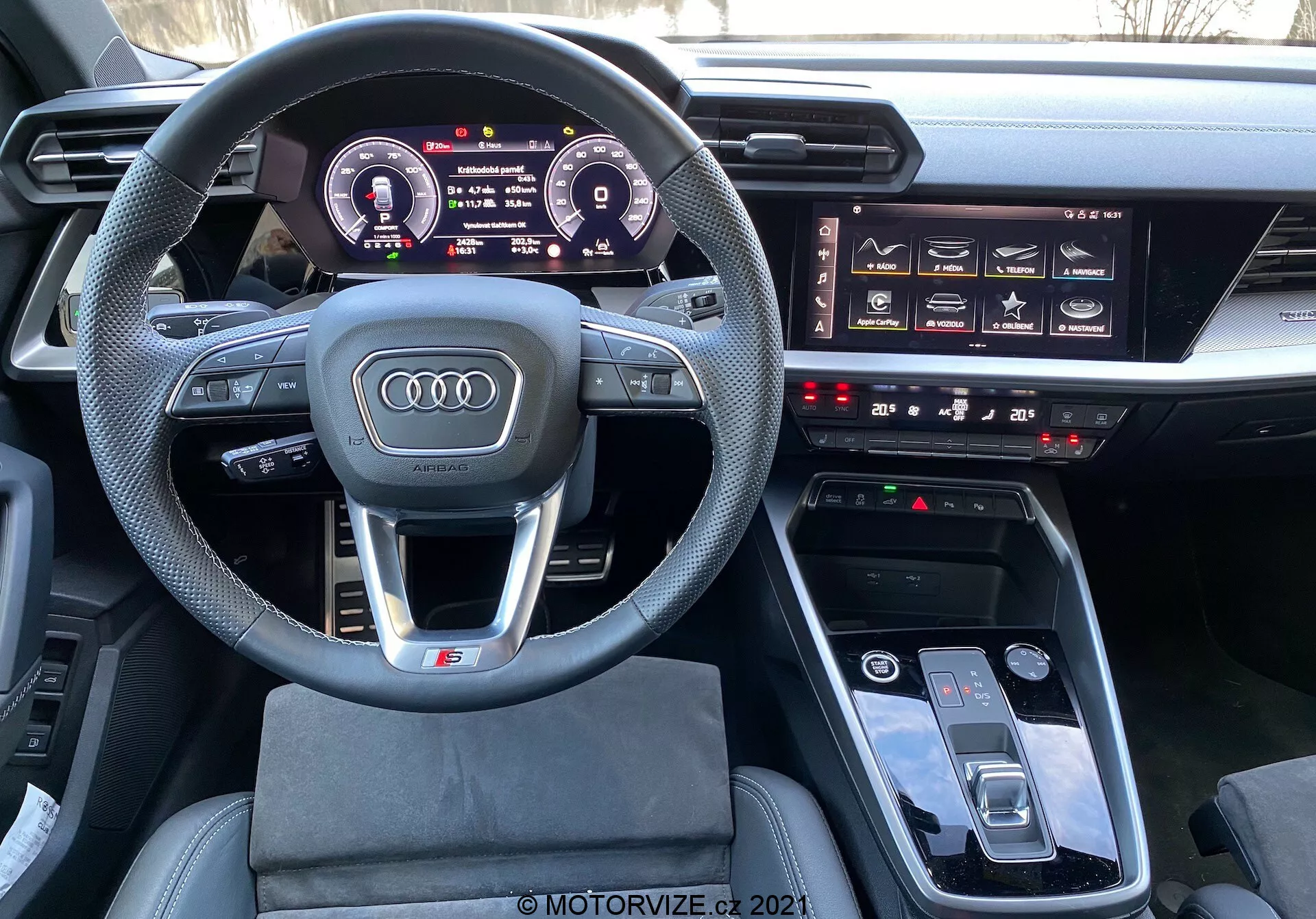 Widok wnętrza Audi A3 Sportback 2020 z perspektywy fotela kierowcy pokazujący wielofunkcyjną, sportową kierownicę obszytą skórą z płaskim spodem, cyfrowy zestaw wskaźników Audi Virtual Cockpit, widoczny system infotainment, dotykowe przyciski do kontroli klimatu i panel dotykowy MMI na konsoli środkowej, wskazujący na nowoczesne, luksusowe i technologicznie zaawansowane wnętrze samochodu.