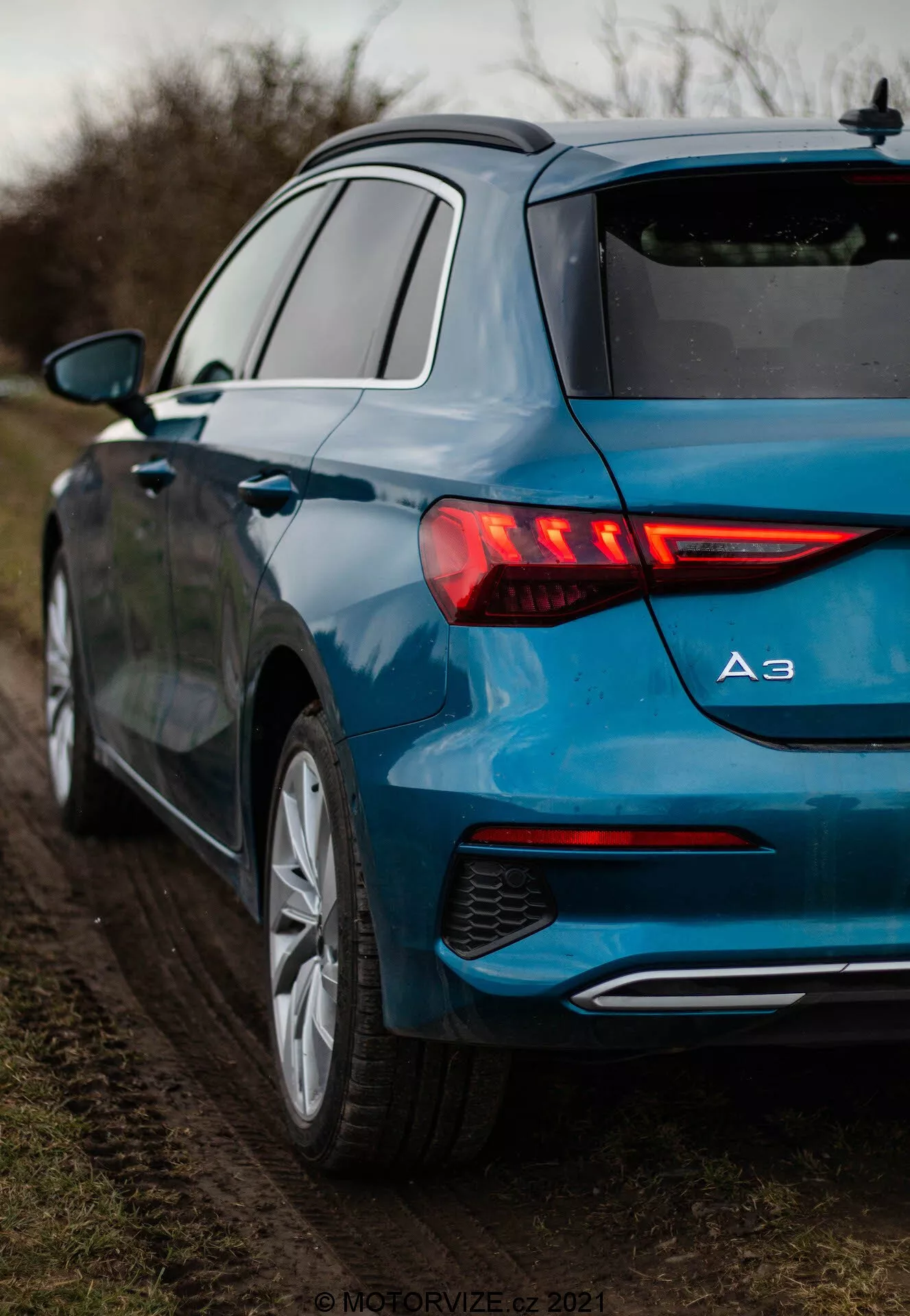 Szczegółowy widok metalicznie niebieskiego Audi A3 2020 Sportback z tylnej perspektywy trzech czwartych, podkreślający jego podświetlane tylne światła LED z dynamicznymi kierunkowskazami, znaczek modelu pokazujący 'A3', wieloramienne alufelgi i pojedynczy wylot wydechowy. Samochód jest zaparkowany na nieutwardzonym poboczu otoczonym roślinnością, w miękkim, rozproszonym oświetleniu.