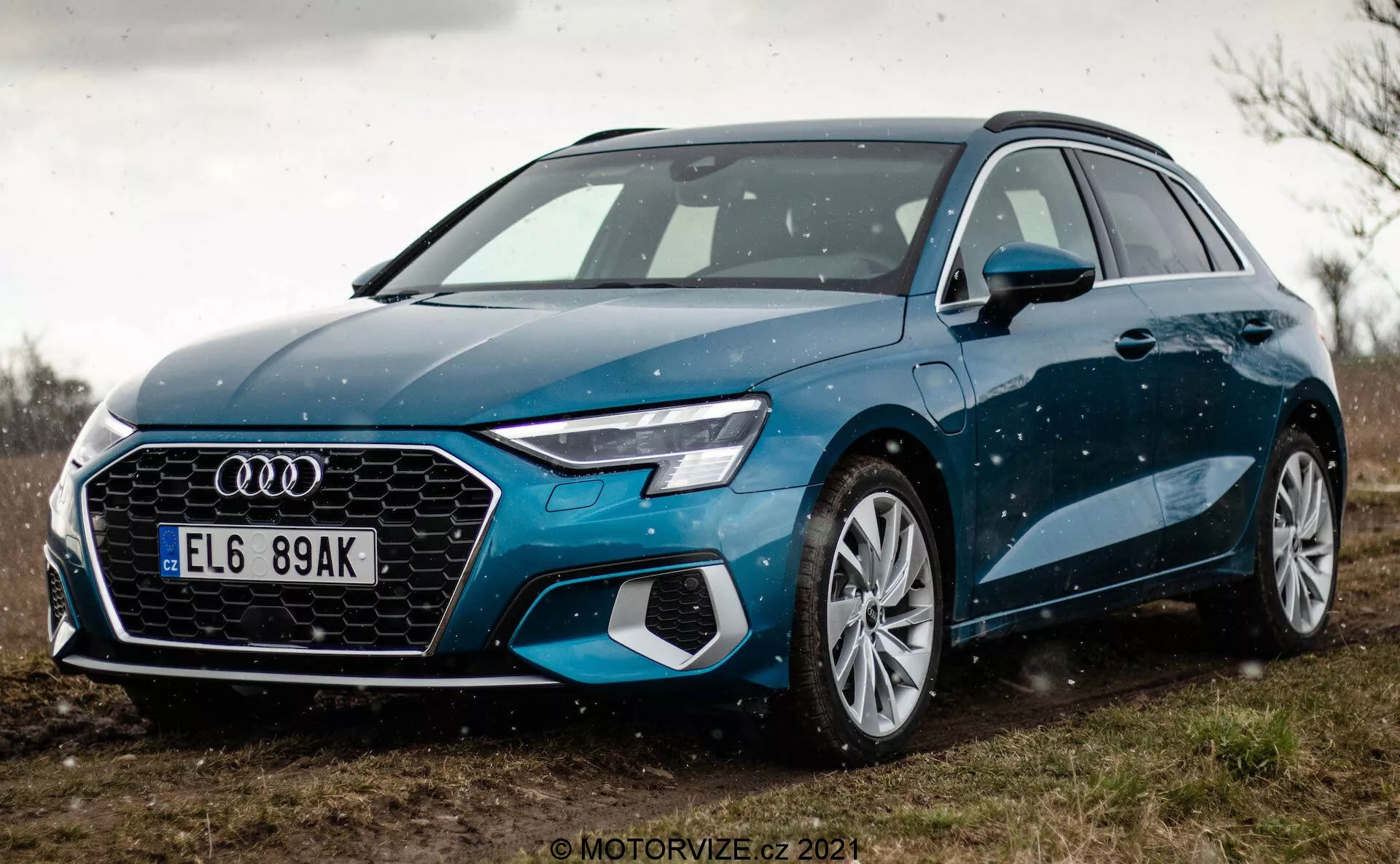 Widok z niskiego kąta na niebieskie Audi A3 Sportback 2020, podkreślający przedni grill i design reflektorów LED. Perspektywa z bliska skupia się na przedniej lewej stronie samochodu, pokazując jego sześciokątną kratkę z wyraźnym emblematem Audi, zintegrowane czujniki funkcji ADAS, duże wieloramienne felgi aluminiowe i eleganckie lusterka boczne. Samochód zaparkowany jest na nieutwardzonej powierzchni z zielenią w tle, sugerując wiejskie otoczenie. Delikatne krople wody na karoserii samochodu podkreślają jego linie i kolor.