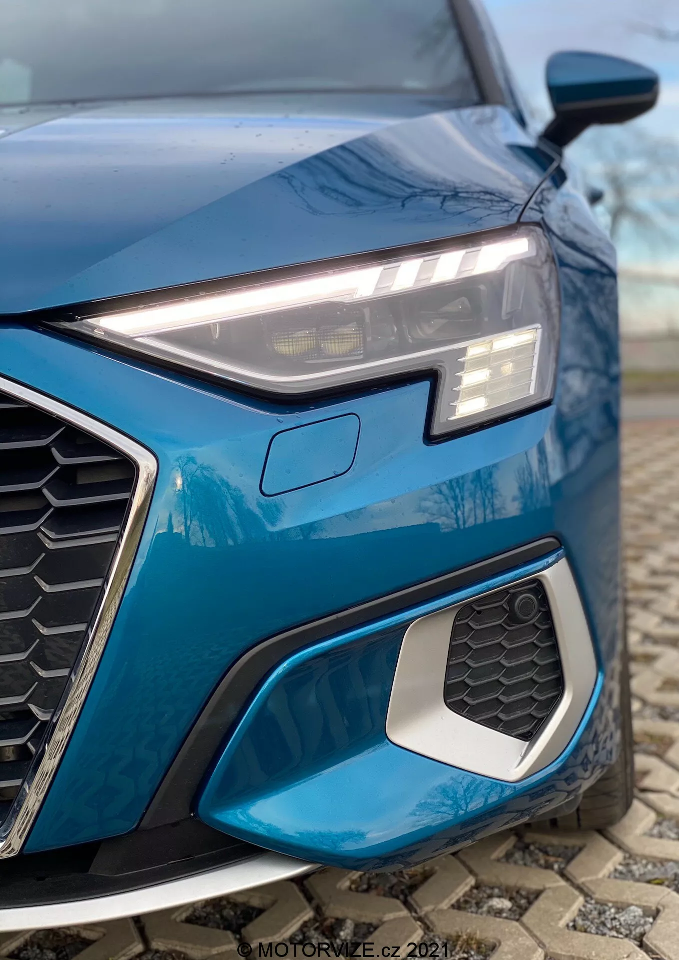 Zbliżenie na przedni lewy kwartał niebieskiego Audi A3 2020 (A3 Sportback), podkreślające elegancki, kanciasty reflektor LED z światłami do jazdy dziennej, wyraźny sześciokątny grill z wzorem plastra miodu oraz wlot powietrza z metalicznymi krawędziami. Nisko kadrowane zdjęcie uwydatnia sportowe elementy designu, złożony geometryczny wzór świateł przeciwmgielnych oraz rozmyte tło zapewniające skupienie na szczegółach.
