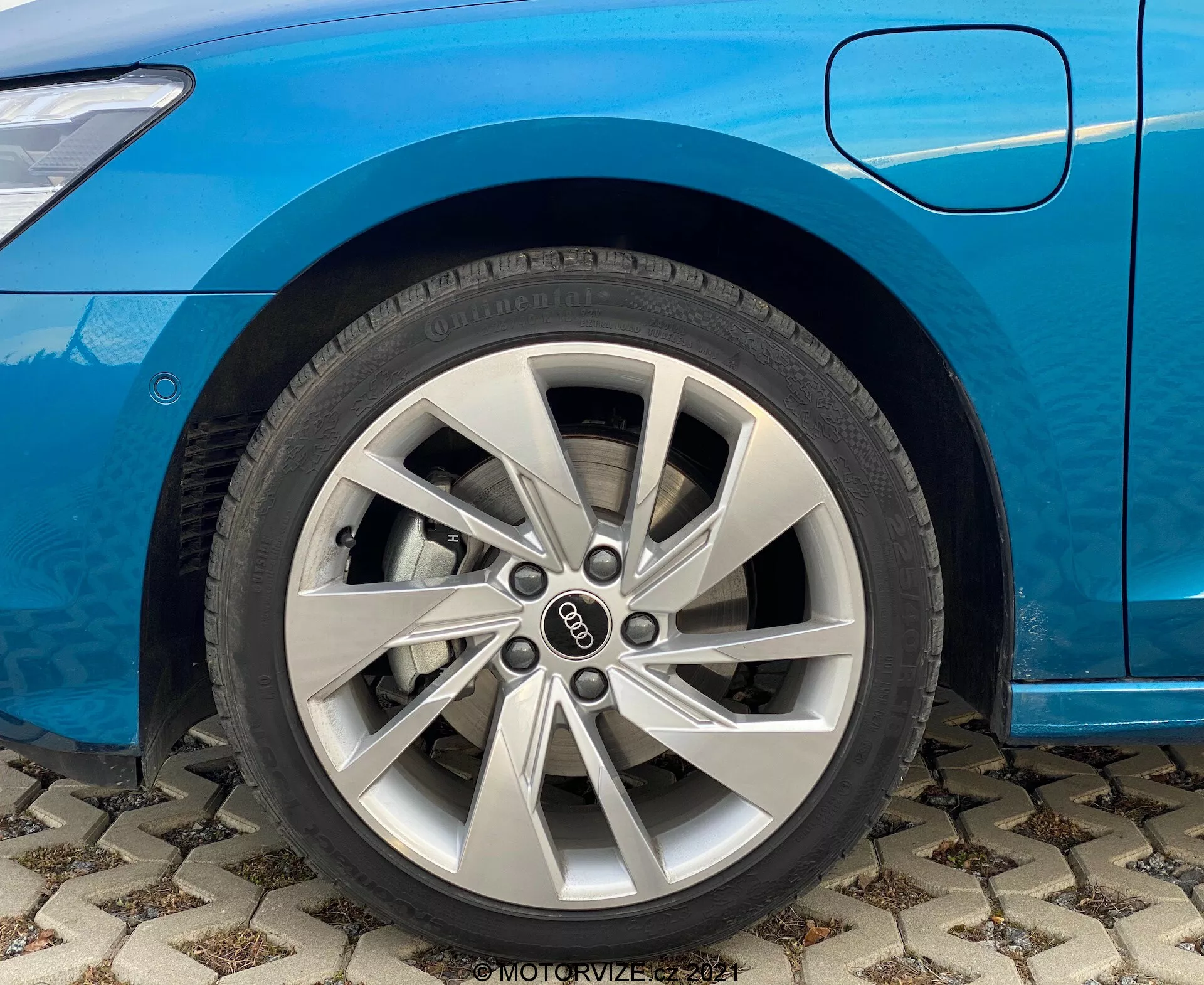 Zbliżenie sekcji przedniego koła niebieskiego Audi A3 2020 (A3 Sportback). Skupiony widok na srebrne wieloramienne koło ze stopu aluminium po stronie pasażera, widoczny również przedni błotnik i dolna krawędź drzwi pasażera. Samochód jest zaparkowany na wzorzystej nawierzchni, z widocznymi detalami bieżnika i oznakowania opony. Istotne dla dyskusji o projektowaniu motoryzacyjnym i technikach produkcji kół. Zawiera znak wodny z napisem 'MOTORVIZE 2021.'