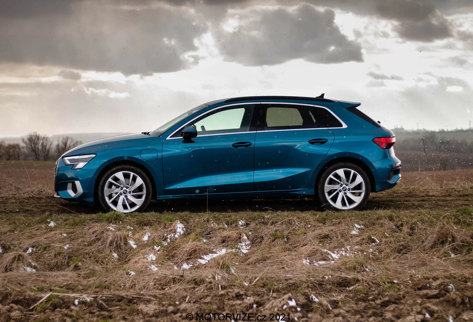 Boczny widok niebieskiego Audi A3 Sportback z 2020 r., zaparkowanego na nierównym trawiastym polu z łatami śniegu lub lodu oraz pochmurnym niebem, podkreślający dynamiczny design samochodu z delikatnie nachylonym dachem, wieloramiennymi felgami aluminiowymi, lekko przyciemnianymi szybami, lusterkami bocznymi w kolorze nadwozia z wbudowanymi kierunkowskazami oraz ostrymi reflektorami LED i tylnymi światłami.
