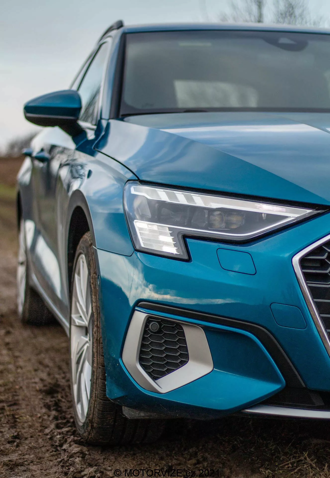 Szczegółowy zdjęcie z ukośnym kątem Audi A3 Sportback 2020, skoncentrowany na lewym przednim kwadrancie. Fotografia z niskiego kąta uchwyca ostre i kanciaste lewy przedni reflektory z LED dziennymi światłami, część sześciokątnej kratki plastra miodu, lewy przedni błotnik i segment drzwi kierowcy. Widoczna jest także felga aluminiowa z wieloramiennym wzorem, metaliczny niebieski lakier i lusterko boczne w kolorze nadwozia z wbudowanymi kierunkowskazami, podkreślając nowoczesny design i cechy technologiczne.