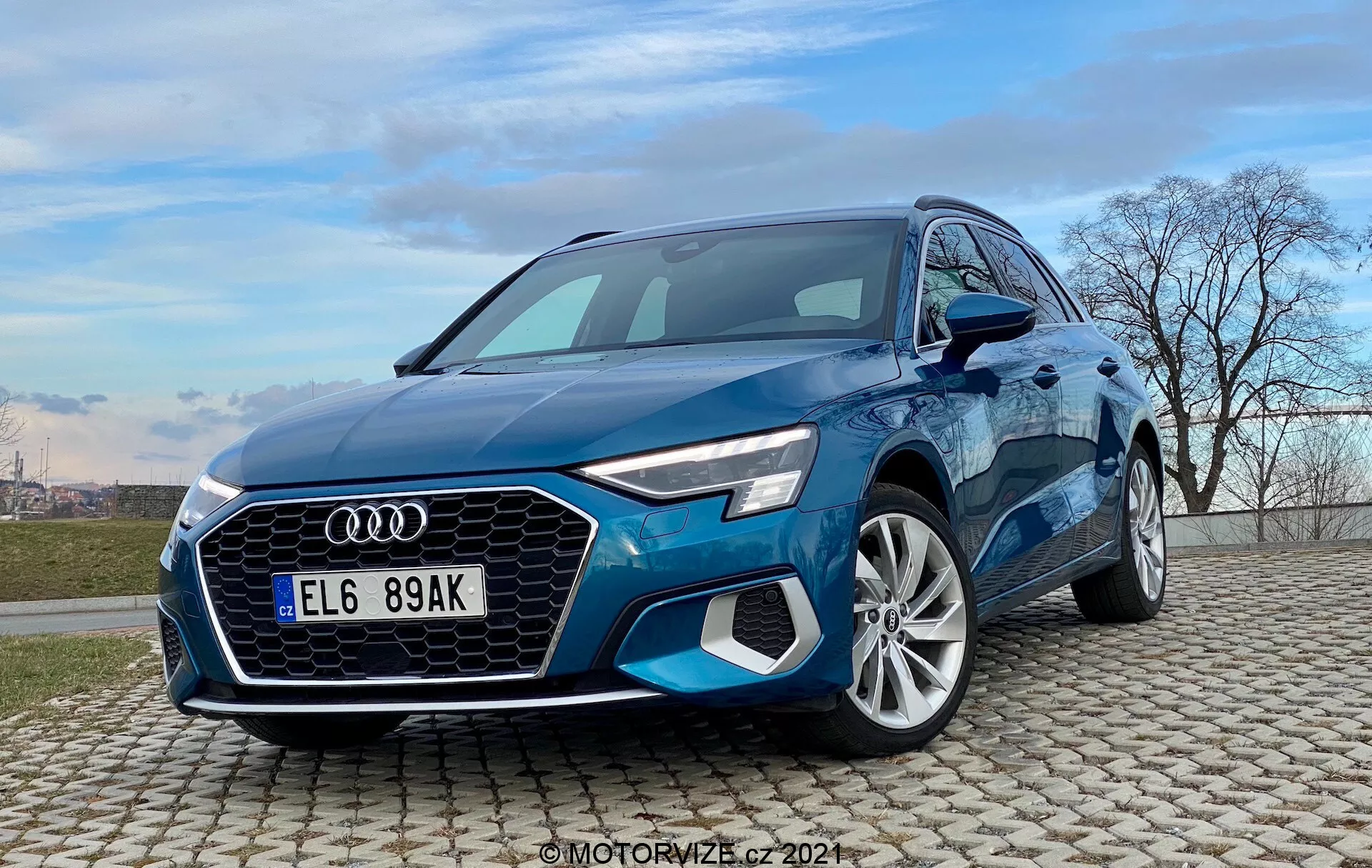 Widok trzy czwarte od przodu niebieskiego Audi A3 Sportback 2020 uchwycony z niskiego kąta. Obraz ukazuje dynamiczny i imponujący wygląd samochodu z charakterystycznym grillem Singleframe, sześciokątnym kształtem z pionowymi żaluzjami, ostrymi reflektorami LED i wlotami powietrza w przednim zderzaku. Rzeźbiona maska prowadzi w stronę przedniej szyby, ujawniając aerodynamiczne kontury i linie nadwozia. Profil boczny przedstawia wyraźnie zarysowane błotniki nad felgami aluminiowymi o wielu szprychach i pochylone słupki A z eleganckimi lusterkami bocznymi. Samochód jest ustawiony na brukowanej nawierzchni pod jasnym, niebieskim niebem z chmurami i linią drzew bez liści, co sugeruje kontekst promocyjny lub recenzji samochodów.