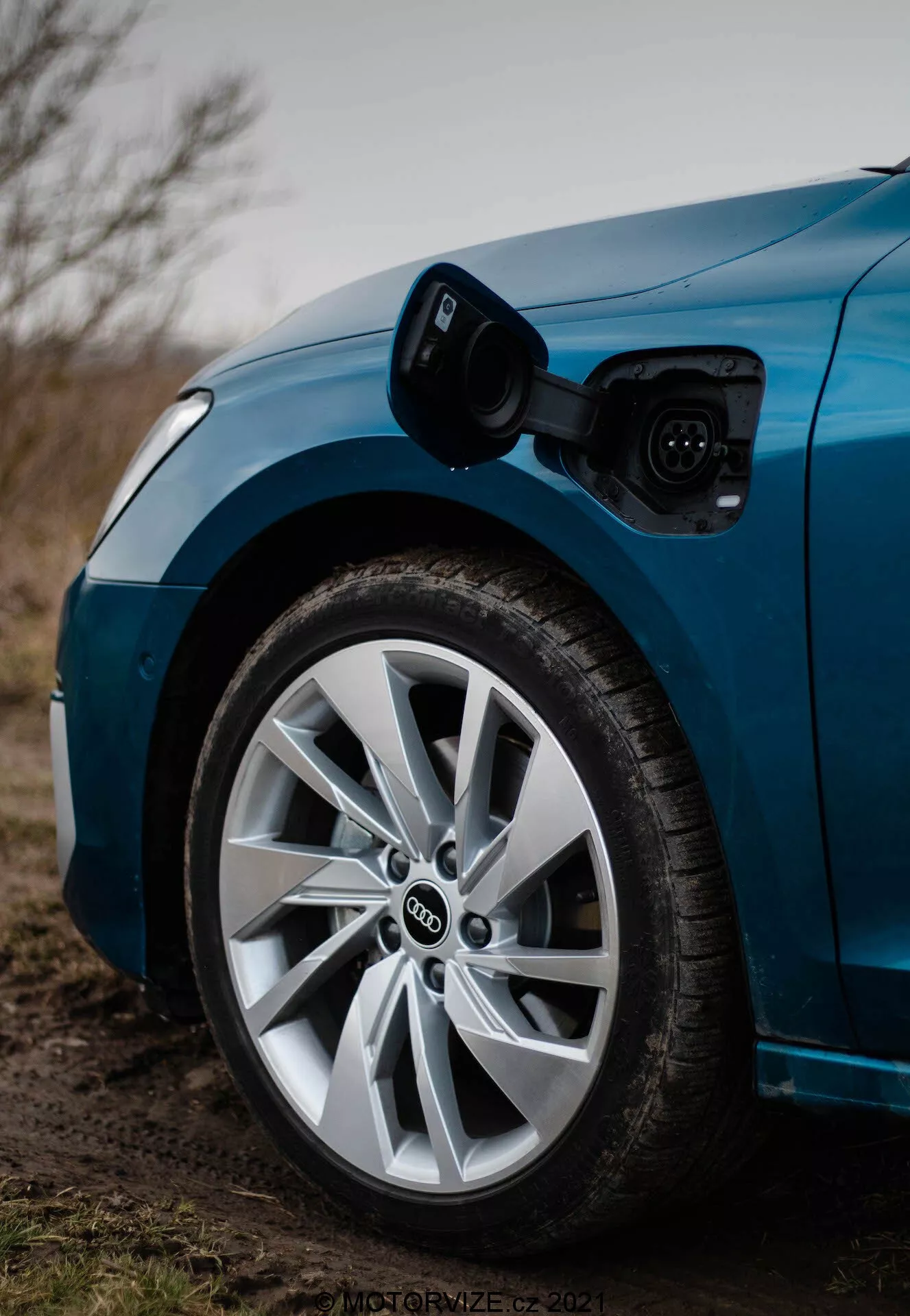 Zbliżenie przedniej części strony kierowcy niebieskiego Audi A3 2020 Sportback, podkreślające koło i port ładowania. Zdjęcie, wykonane z niskiego kąta w kierunku przodu, pokazuje otwarty port ładowania nad przednim kołem, z podłączonym kablem ładującym. Płytka głębia ostrości koncentruje się wyraźnie na kole i porcie ładowania, ukazując charakterystyczne logo Audi składające się z nachodzących na siebie pierścieni oraz wieloszprychowy design koła z niskoprofilową oponą, wszystko w lekko przygaszonym, możliwie zmierzchowym oświetleniu.