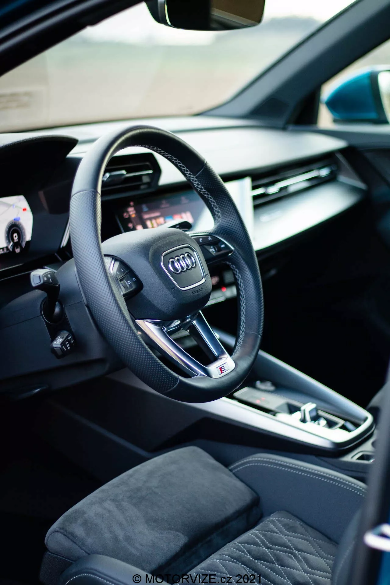 Widok wnętrza Audi A3 Sportback 2020, skoncentrowany na stronie kierowcy z nowoczesną, trójramienną kierownicą z logo Audi, zintegrowanymi przyciskami wielofunkcyjnymi i płaskim dołem, co wskazuje na sportowe osiągi. Za kierownicą widoczna jest część cyfrowego zestawu wskaźników lub wirtualnego kokpitu, oferującego konfigurowalne opcje wyświetlania dla różnych funkcji samochodu. Po lewej stronie otwarte drzwi samochodu ukazują sterowanie elektrycznymi szybami i ustawieniami pamięci fotela, podkreślając wygodę i personalizację. Konsola środkowa obejmuje automatyczny wybierak biegów, przycisk elektronicznego hamulca postojowego i panel dotykowy/sterownik systemu multimedialnego, podkreślając zaangażowanie kierowcy. Widoczne fotele są tapicerowane wysokiej jakości materiałami z pikowanym wzorem, dodającym wnętrzu luksusowego charakteru.