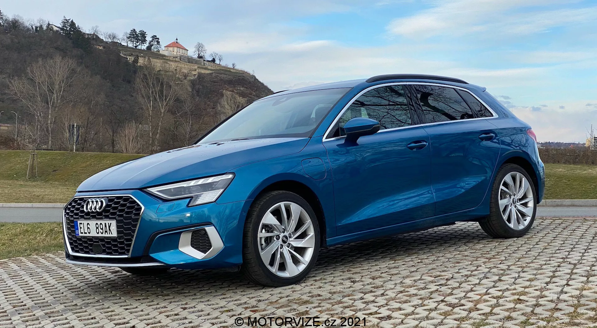 Widok z przodu pod kątem trzy czwarte jasnoniebieskiego Audi A3 Sportback 2020 na bruku, pokazujący charakterystyczne elementy designu, takie jak reflektory LED, duża krata, wieloramienne felgi aluminiowe i zaawansowane systemy bezpieczeństwa. Ujęcie z tłem przypominającym pagórkowaty teren z trawą oraz częściowo zachmurzone niebo.