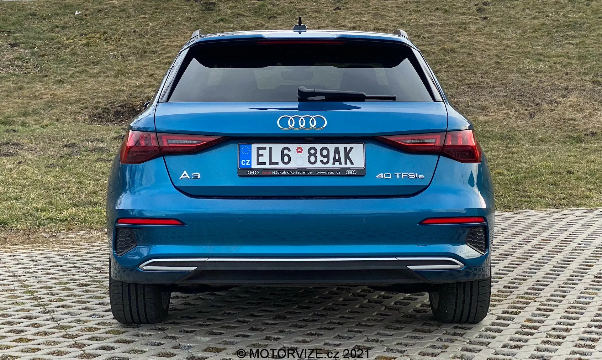 Widok z tyłu niebieskiego Audi A3 2020 Sportback, ukazujący tylny zderzak, bagażnik, tylne światła, rury wydechowe oraz tylną szybę. Wyraźne logo Audi i oznaczenie modelu 'A3' z plakietką '40 TFSI' wskazującą na turbodoładowany silnik benzynowy. Tylne światła LED o eleganckim poziomym designie, podwójne rury wydechowe, tylny zderzak z elementem dyfuzora i czujnikami parkowania oraz tablica rejestracyjna w europejskim stylu. Tło stanowi trawiasta skarpa.