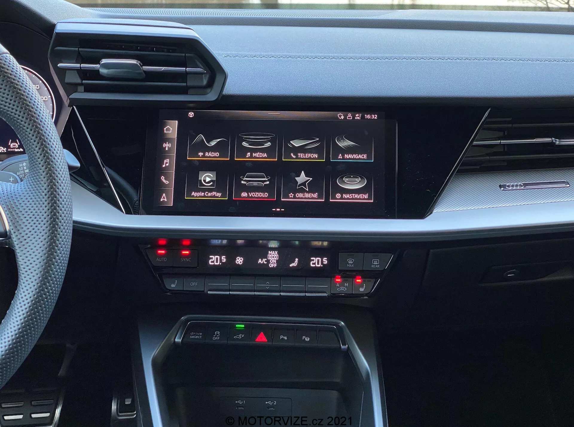 Widok wnętrza deski rozdzielczej Audi A3 Sportback 2020, podkreślający zaawansowany system informacyjno-rozrywkowy oraz sterowanie klimatyzacją. Obraz uchwyca duży poziomy ekran dotykowy z funkcjami takimi jak nawigacja, media, łączność telefoniczna i ustawienia samochodu, wskazując na kompleksowe możliwości infotainment w tym Apple CarPlay i Android Auto. Poniżej ekranu znajdują się cyfrowe sterowniki klimatyzacji dwustrefowej, pokazujące ustawienia na 20 stopni Celsjusza dla kierowcy i pasażera. Deska rozdzielcza zawiera również obszar do ładowania bezprzewodowego oraz cienkie, poziome otwory wentylacyjne, ilustrujące nowoczesny design wnętrza samochodowego. Zdjęcie jest oznaczone znakiem wodnym 'MOTORVIZE.CZ 2021'.