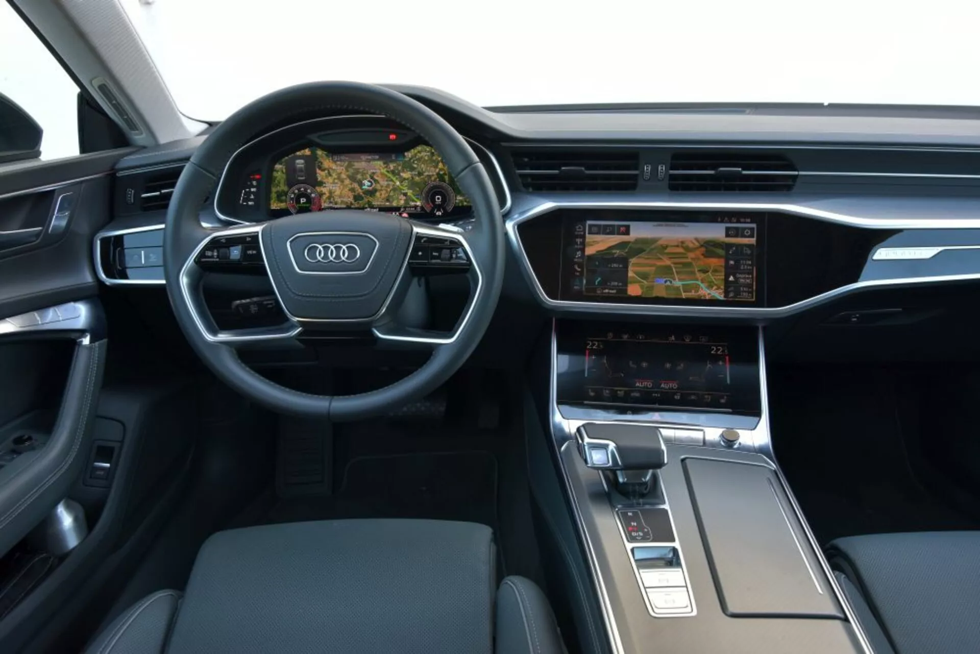 Wnętrze Audi A7 Sportback 2018 skupiające się na desce rozdzielczej kierowcy i obszarze konsoli środkowej, prezentujące wielofunkcyjną kierownicę z trzema ramionami z logo Audi, cyfrowy zestaw wskaźników Virtual Cockpit wyświetlający nawigację, system infotainment z podwójnym wyświetlaczem z ustawieniami klimatyzacji, ergonomiczny selektor biegów i materiały premium takie jak szczotkowane metal i skóra.