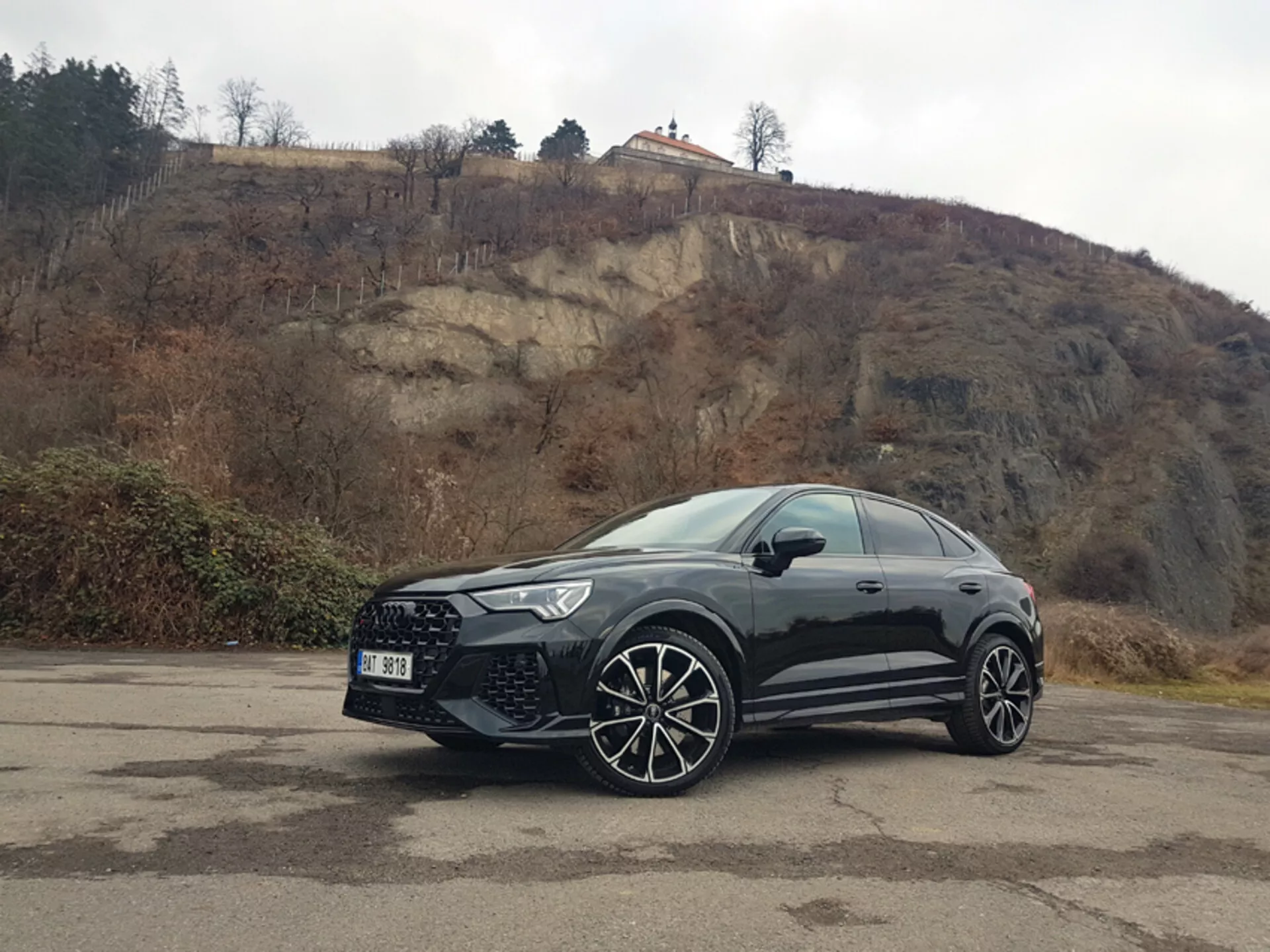 Boczny przedni widok Audi RS-Q3 z 2015 roku zaparkowanego na zewnątrz na wyblakłym parkingu z kostki brukowej, prezentujący felgi aluminiowe i przedni grill na tle pochmurnego wzgórza
