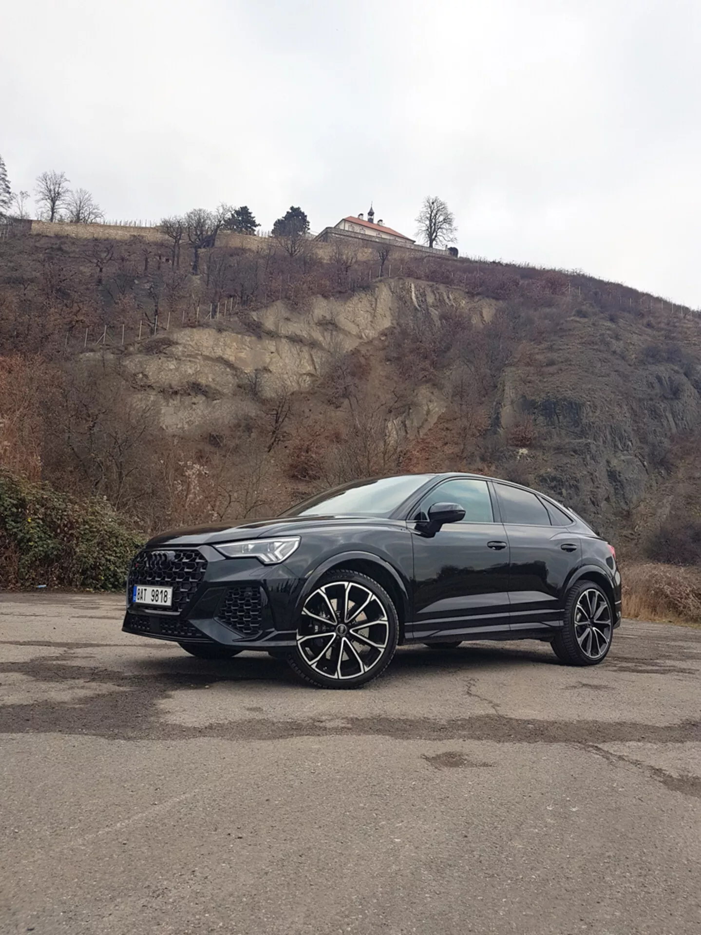 Czarny sportowy samochód użytkowy Audi Q3 RS 2015 zaparkowany na wybrukowanym terenie z tłem skalistego wzgórza, prezentujący duże koła z wieloramiennej alufelgi, smukłe reflektory wyposażone w technologię LED i sportowe linie nadwozia pod zachmurzonym niebem.