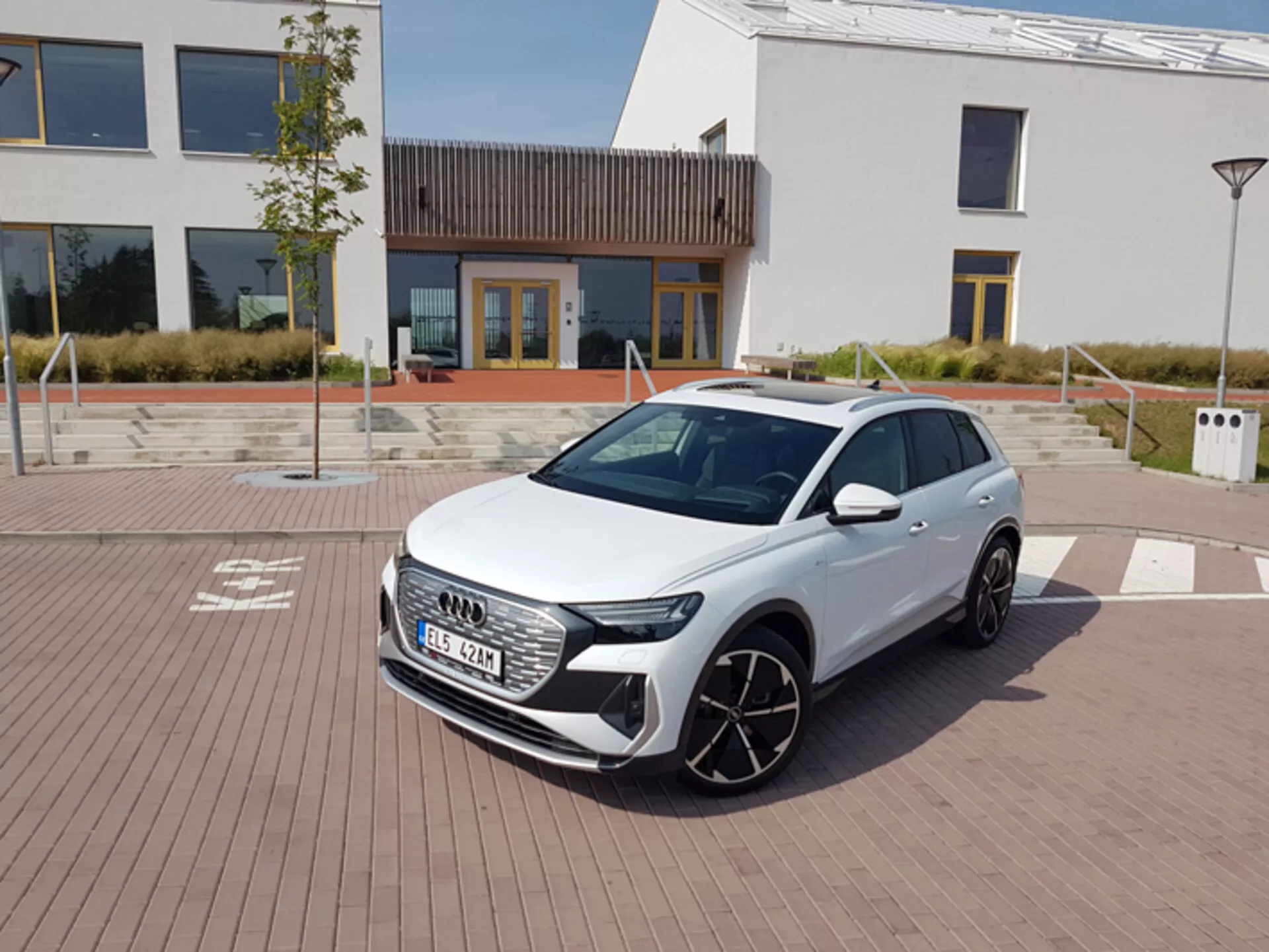 Biały Audi Q4 e-tron 2021, prezentujący nowoczesne reflektory LED, sześciokątną atrapę chłodnicy i duże felgi aluminiowe, zaparkowany na brukowanej powierzchni z wzorem z cegieł z miejską architekturą i stacją ładowania w tle, pod jasnym niebem w południe.