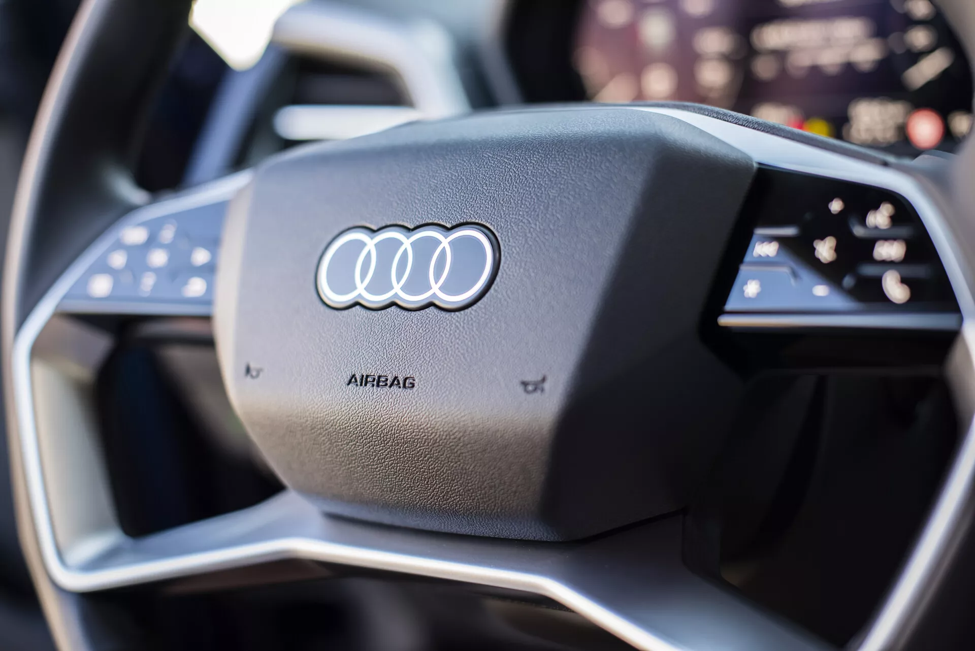 Zbliżenie na kierownicę Audi Q4 e-tron 2021 z ikonicznym logo Audi na piastę poduszki powietrznej, otoczone multifunkcyjnymi przyciskami, z nowoczesnym, sportowym designem obejmującym kontrastowe szwy i sugestią zaawansowanej technologii cyfrowego deski rozdzielczej w tle